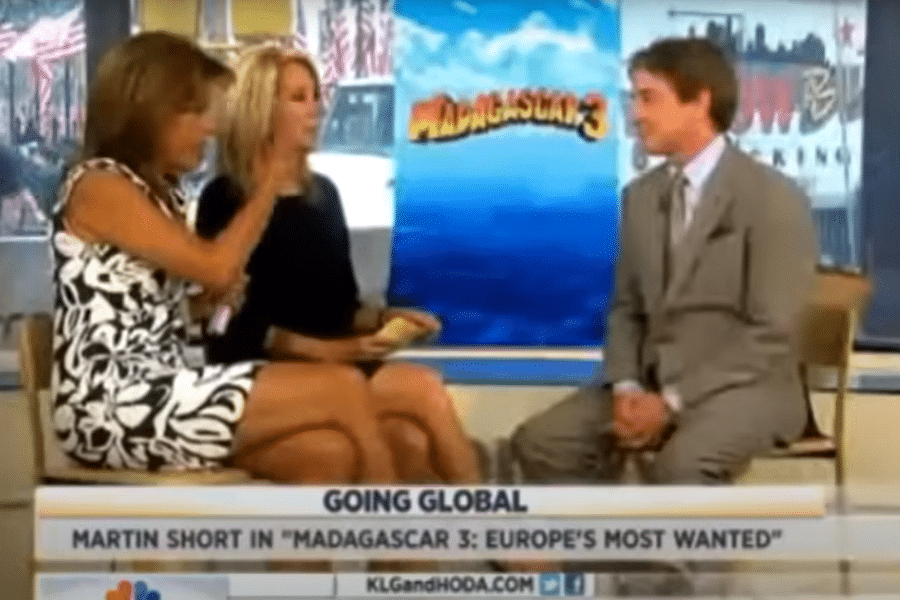 Two women sit on a studio set interviewing a man in a suit. Behind them is a poster for "Madagascar 3: Europe's Most Wanted." The mood is lively.