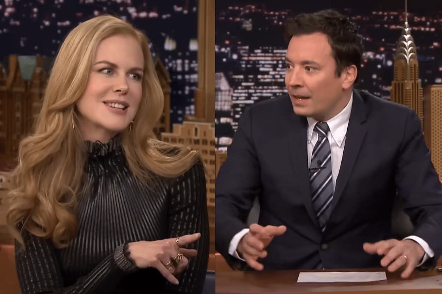 TV talk show scene with a woman in a black dress and a man in a suit. They appear engaged in animated conversation, set against a cityscape backdrop.