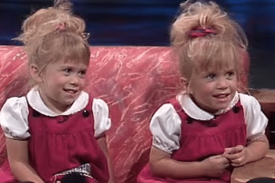Two young twin girls sit side by side on a red patterned couch, smiling while wearing matching pink pinafore dresses over white blouses with their blonde hair styled in high ponytails.