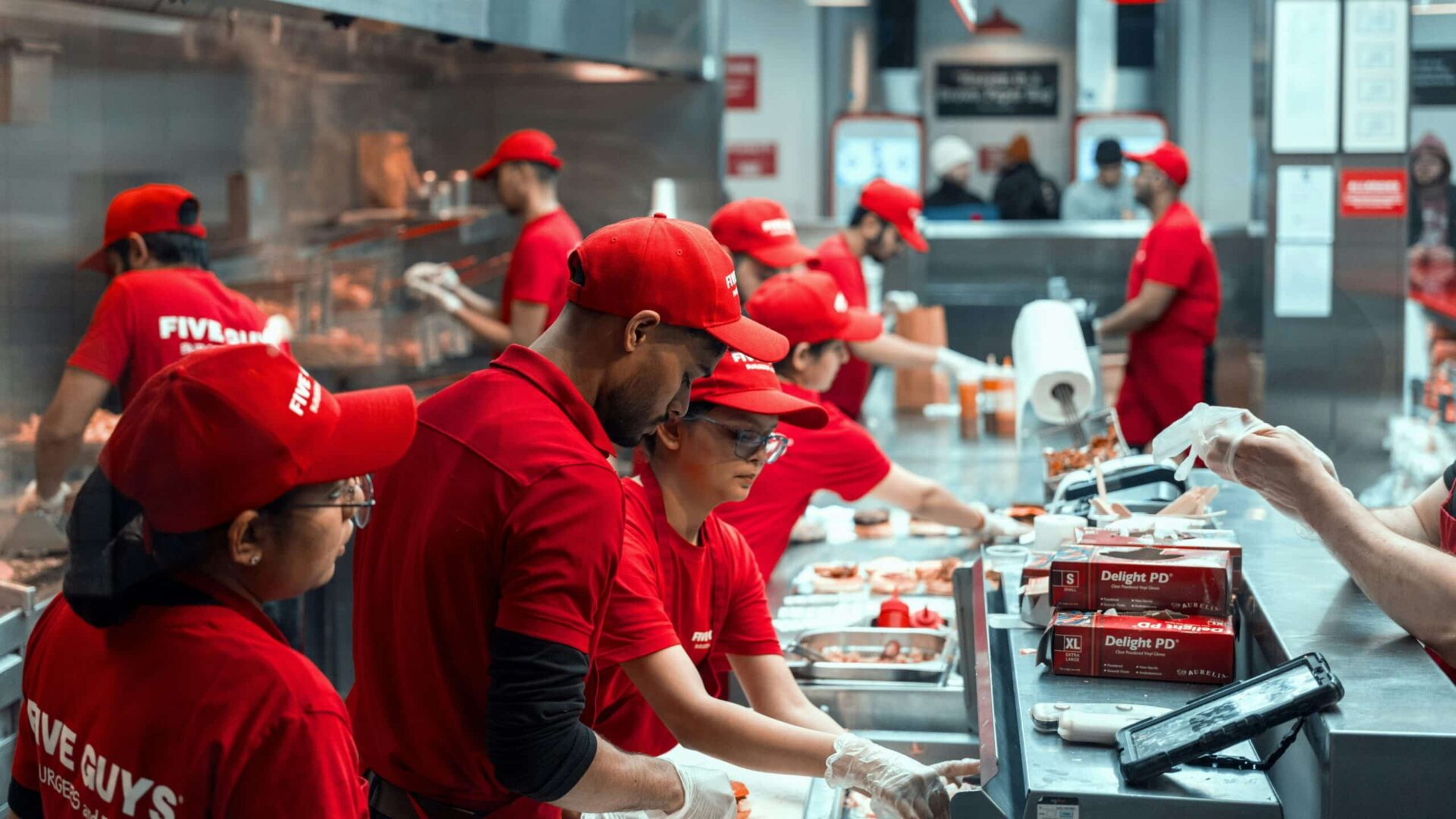 Fast-food employees in red uniforms preparing food inside a restaurant kitchen.