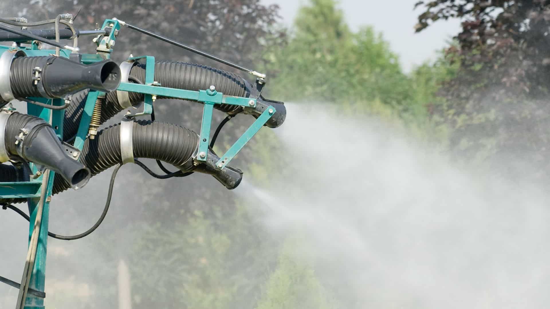 Industrial sprayer nozzles mounted on a green frame emitting a thick, white mist or pesticide over a field of trees.