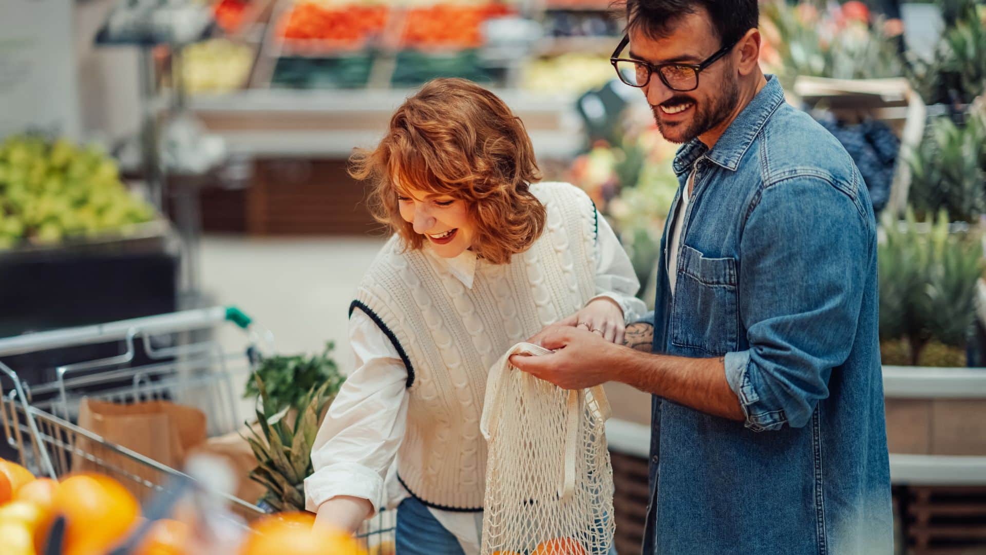 Young couple selecting fresh oranges at a market stall using reusable bags