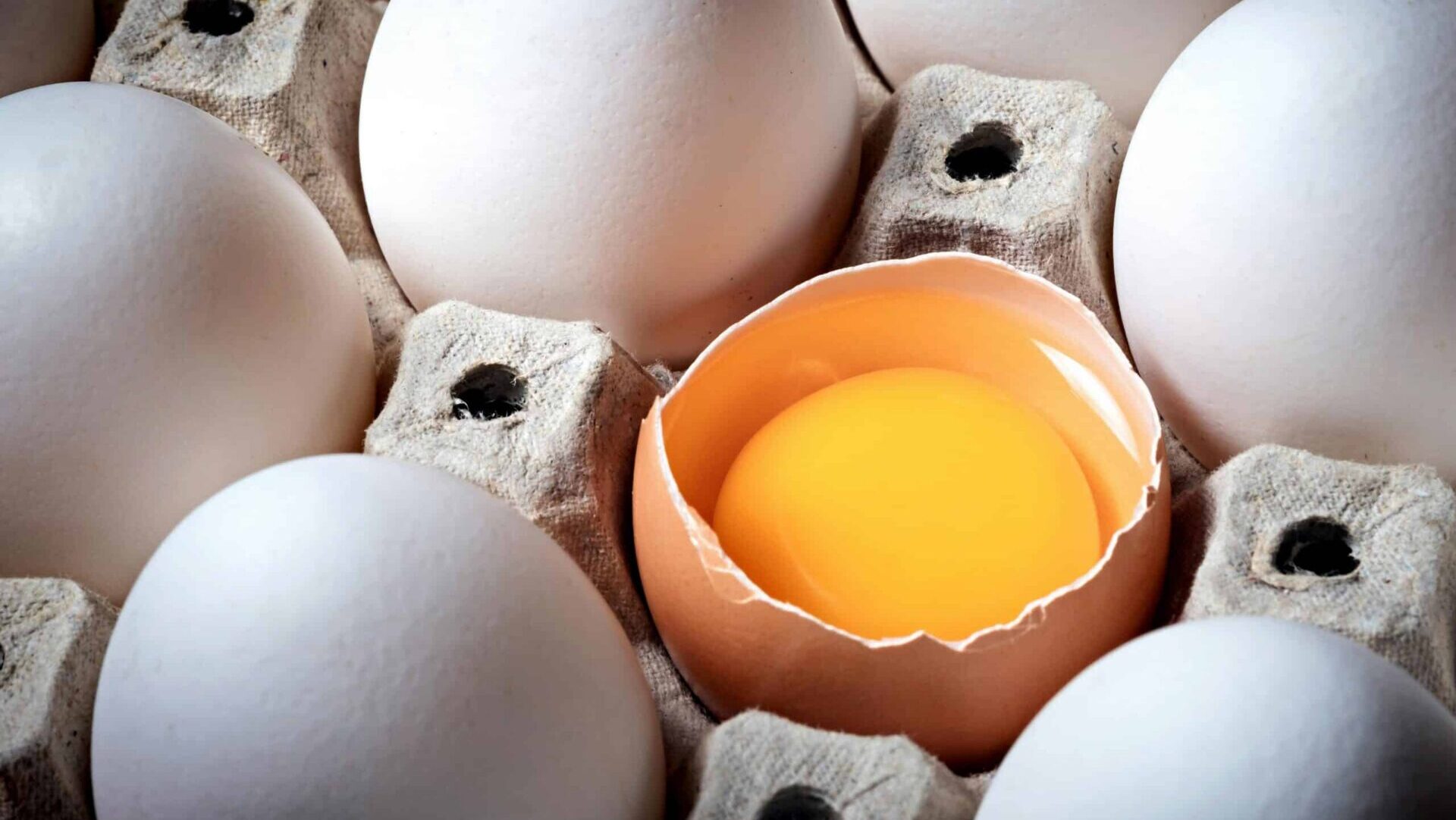 Single broken egg among whole eggs in a carton, showing cracked shell and exposed yolk.