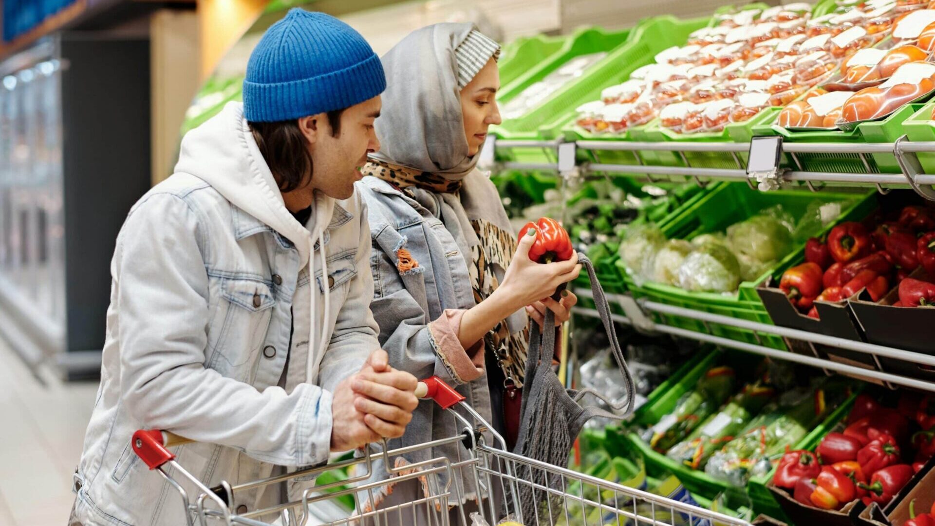 Couple buying groceries in a supermarket aisle with a cart partially filled with food items.