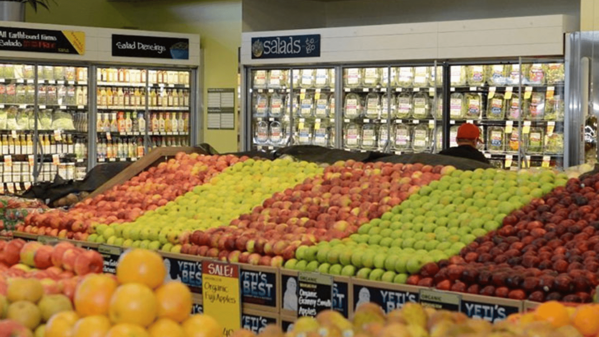 A produce section in a grocery store with neatly stacked apples and other fresh fruits under bright lighting.