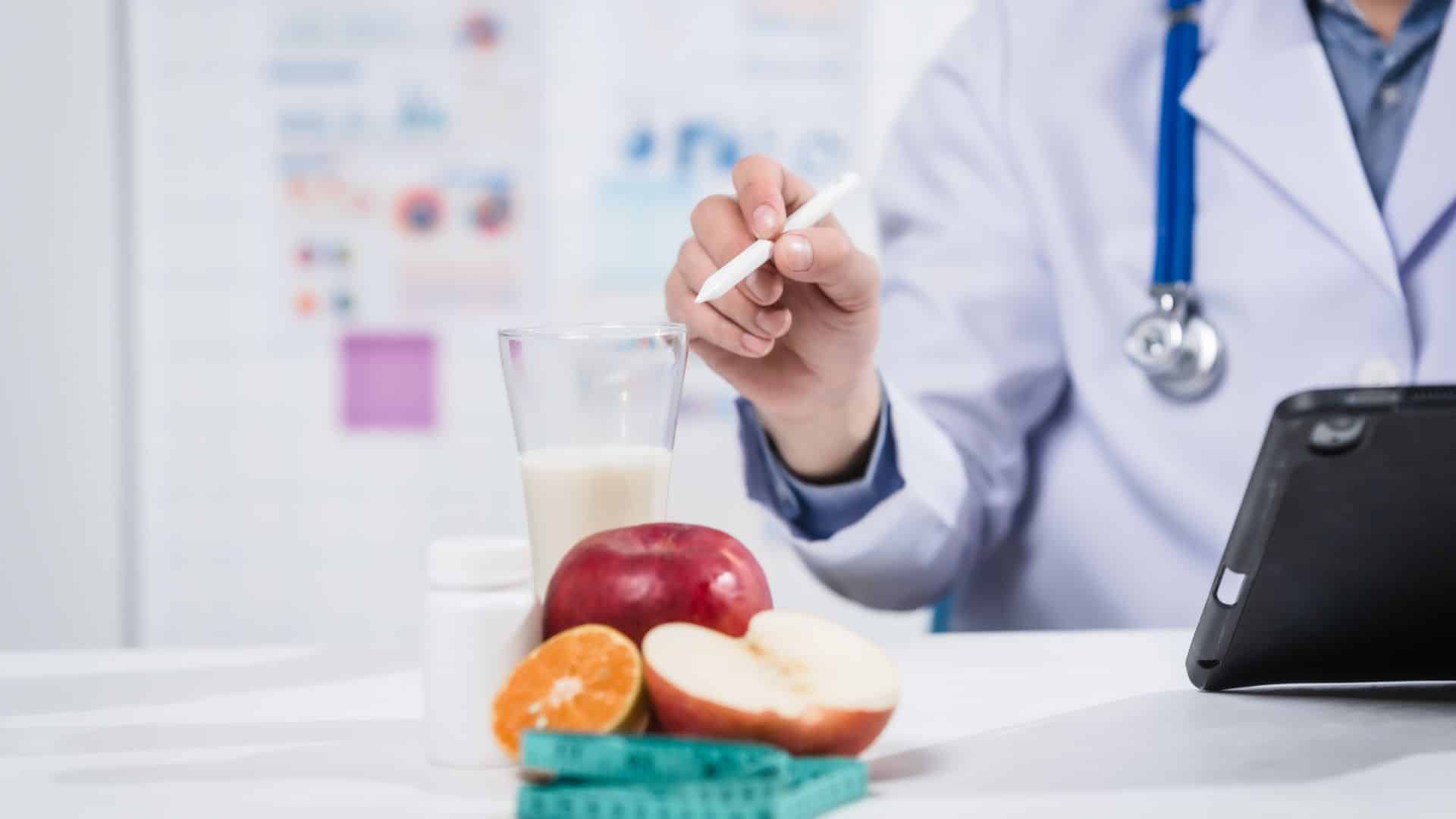 Doctor holding a pen above a desk with fruit, milk, and a measuring tape during a nutrition consultation.