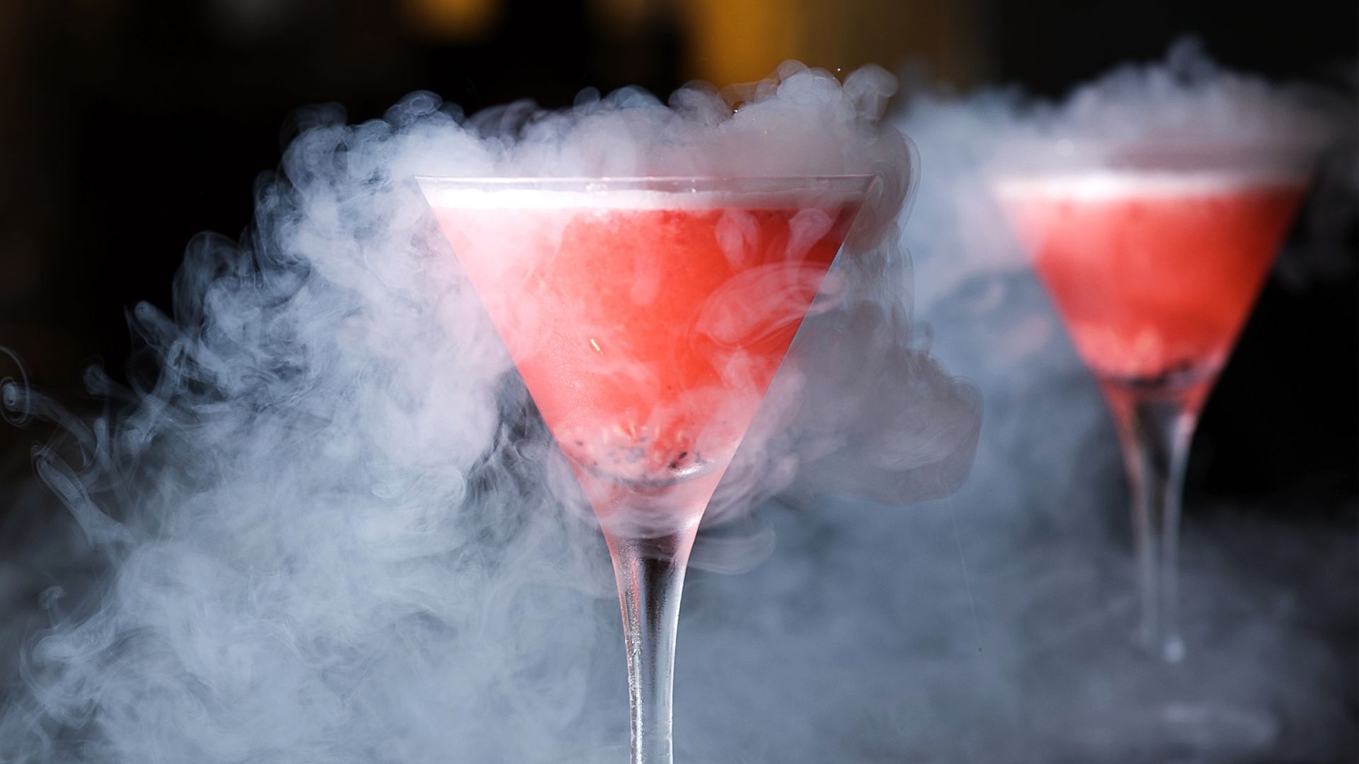 Bright pink cocktail in a martini glass surrounded by thick white vapor created by liquid nitrogen, with a second matching drink softly blurred in the background.