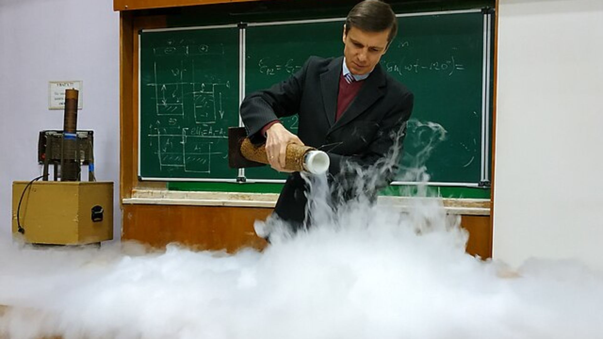 Man in a suit pours liquid nitrogen onto a table during a classroom science demonstration, creating a large cloud of white vapor that spreads across the surface.