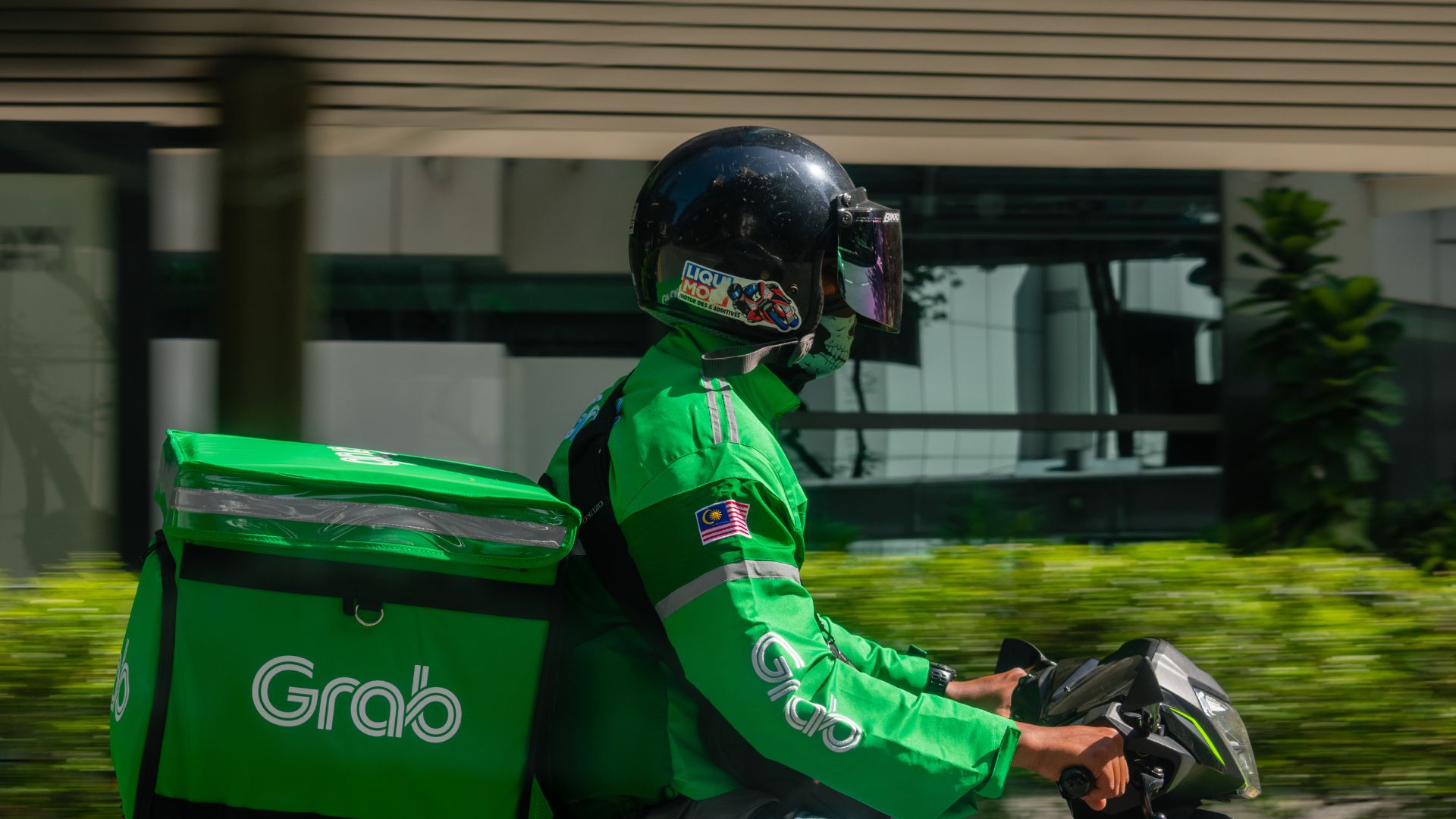 Food delivery rider wearing a green Grab uniform and helmet riding a motorcycle in a city.