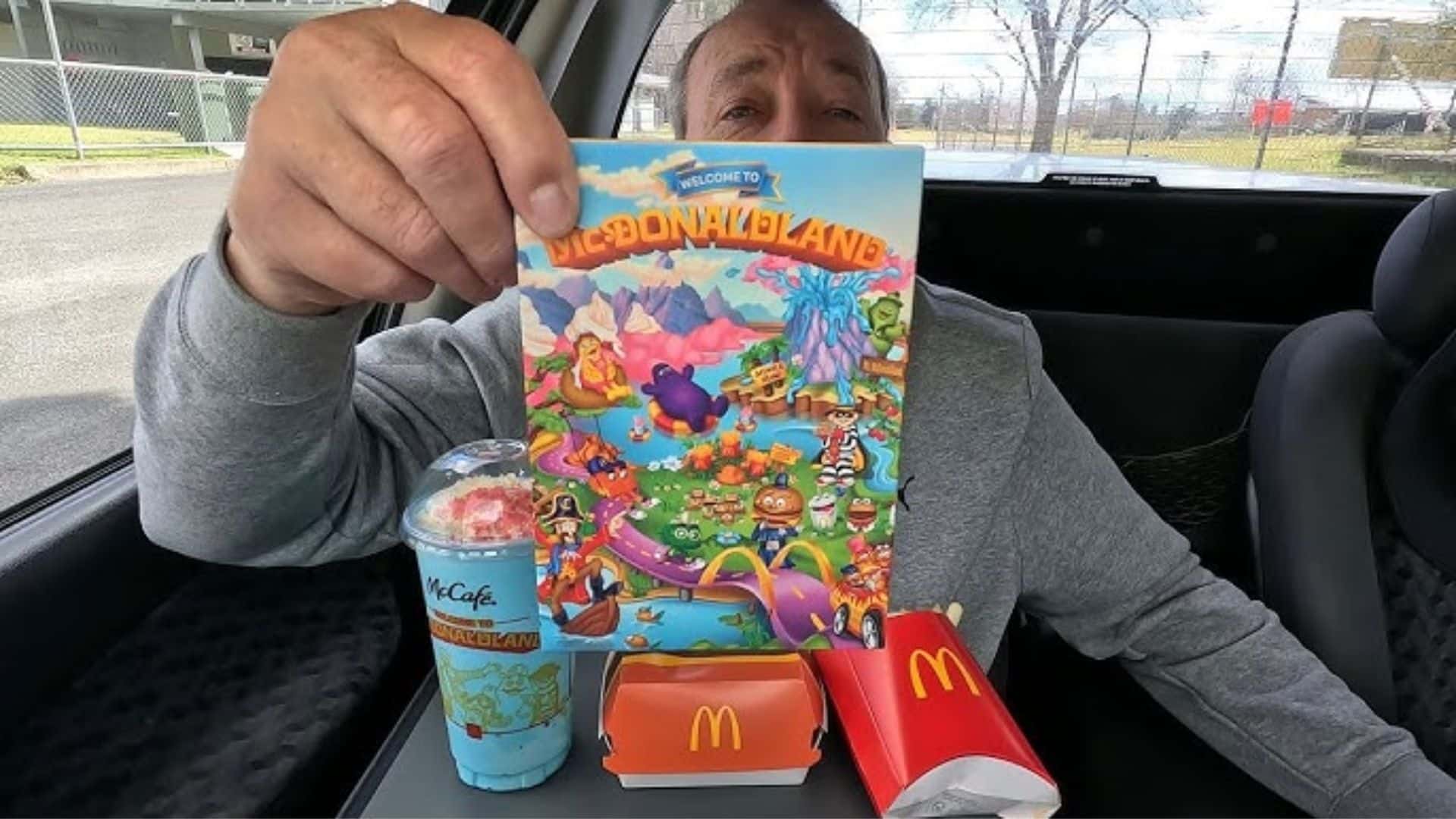 A person sitting in a car holds up a colorful McDonaldland themed card while McDonald’s food and a McCafé drink rest on the center.