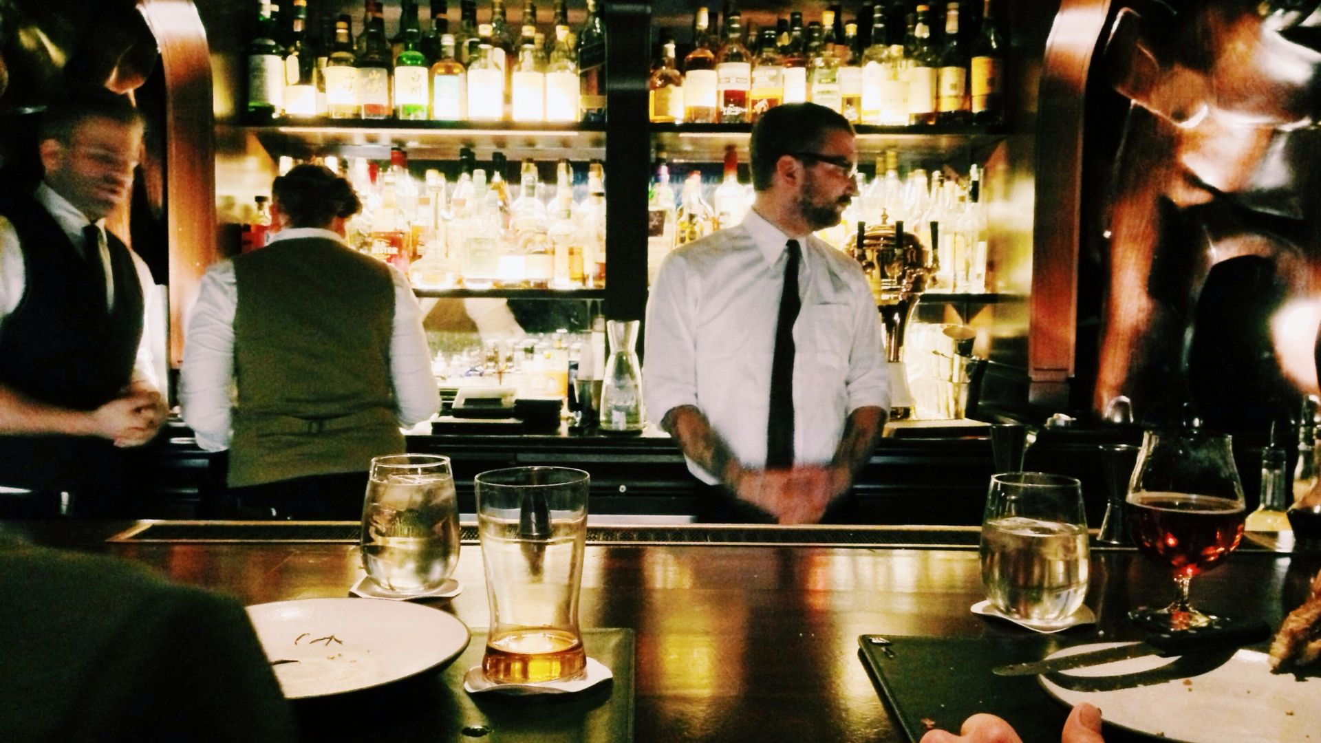 Bartender in a white shirt and black tie stands behind a warmly lit bar lined with liquor bottles while preparing drinks.