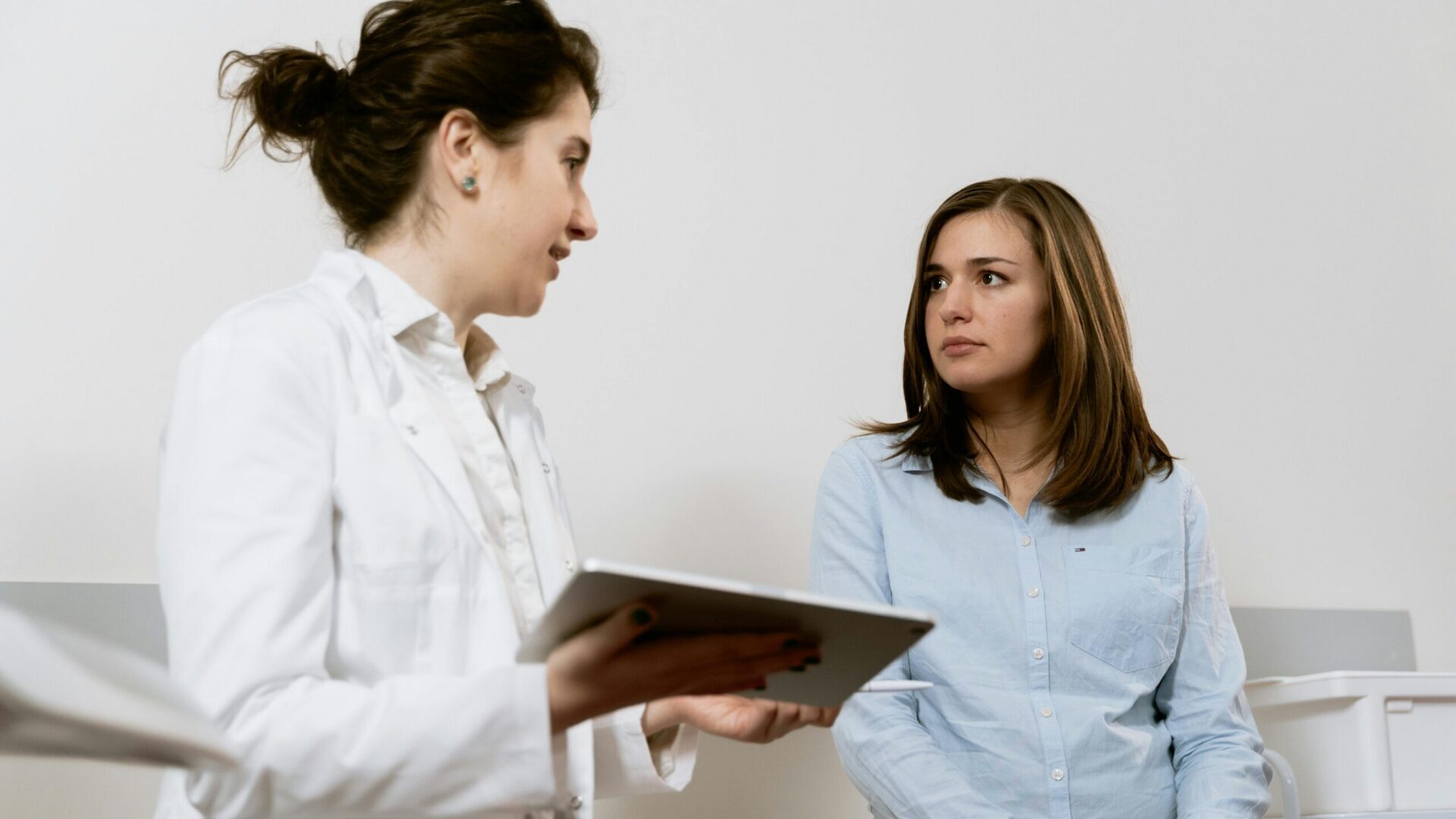 Doctor holding a tablet while speaking with a patient seated in an exam room.