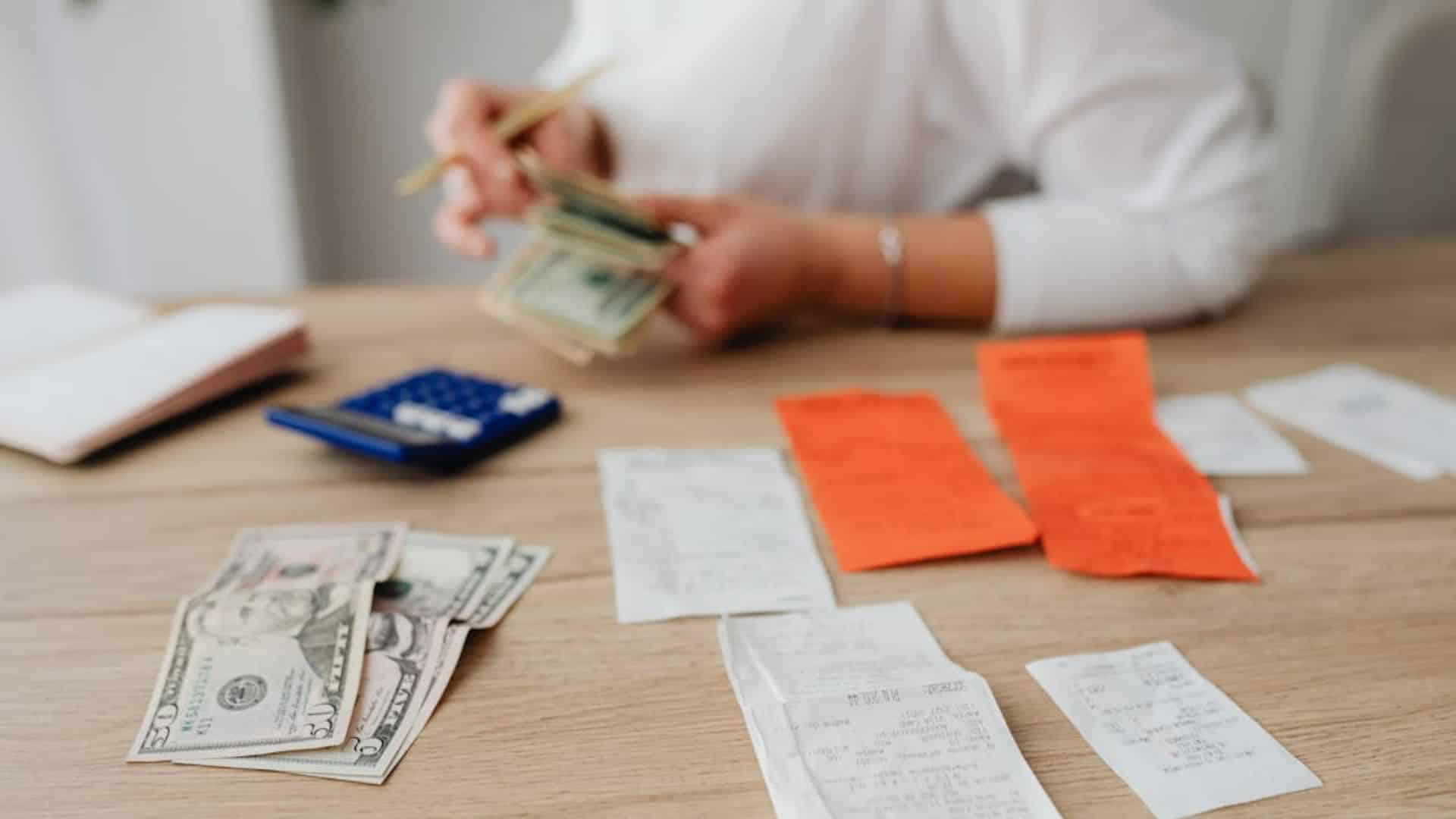 A person counting money at a table with a calculator, receipts, and dollar bills
