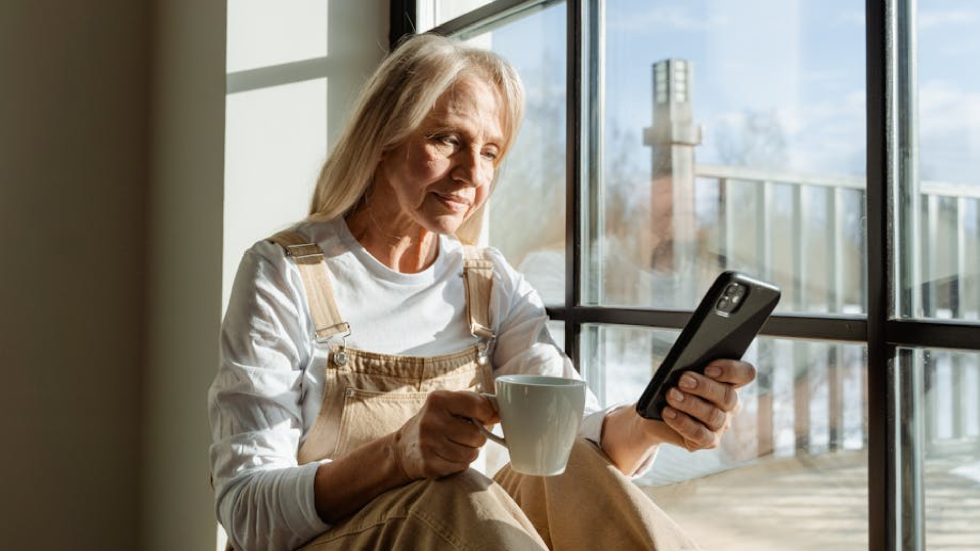 Older woman sitting by a window holding a coffee cup and looking at her smartphone.