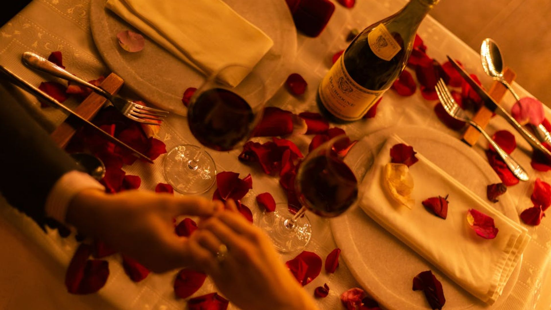 A couple holding hands over a romantically set up table with roses and a bottle of wine
