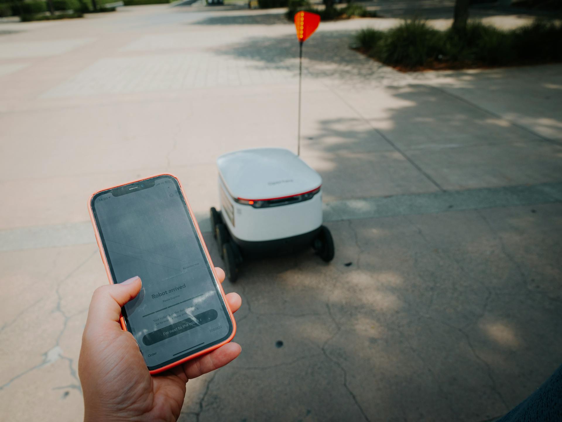A person holding a smartphone displaying a “robot arrived” notification as a food delivery robot waits on the sidewalk.