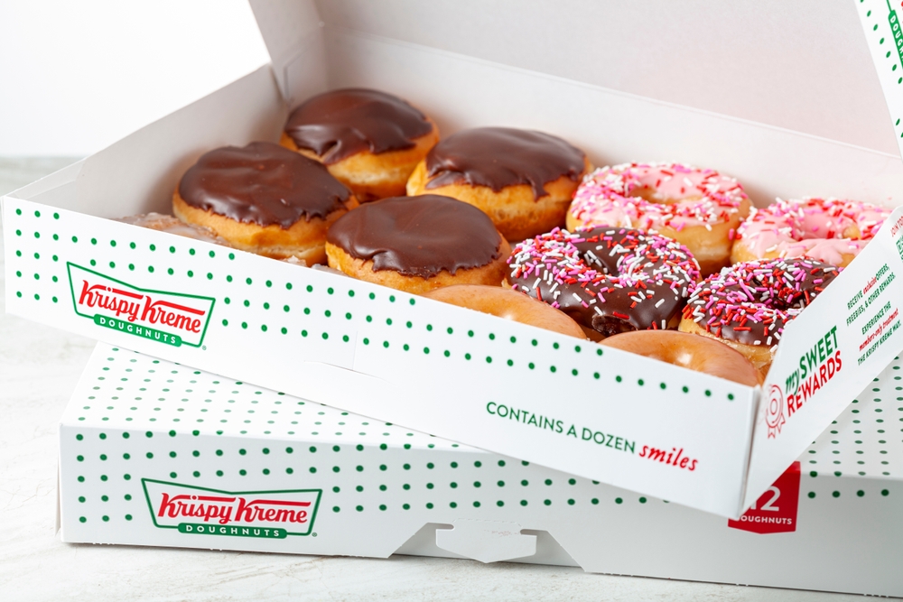 Open Krispy Kreme box filled with assorted doughnuts, including chocolate-glazed and sprinkled varieties, stacked on branded packaging.