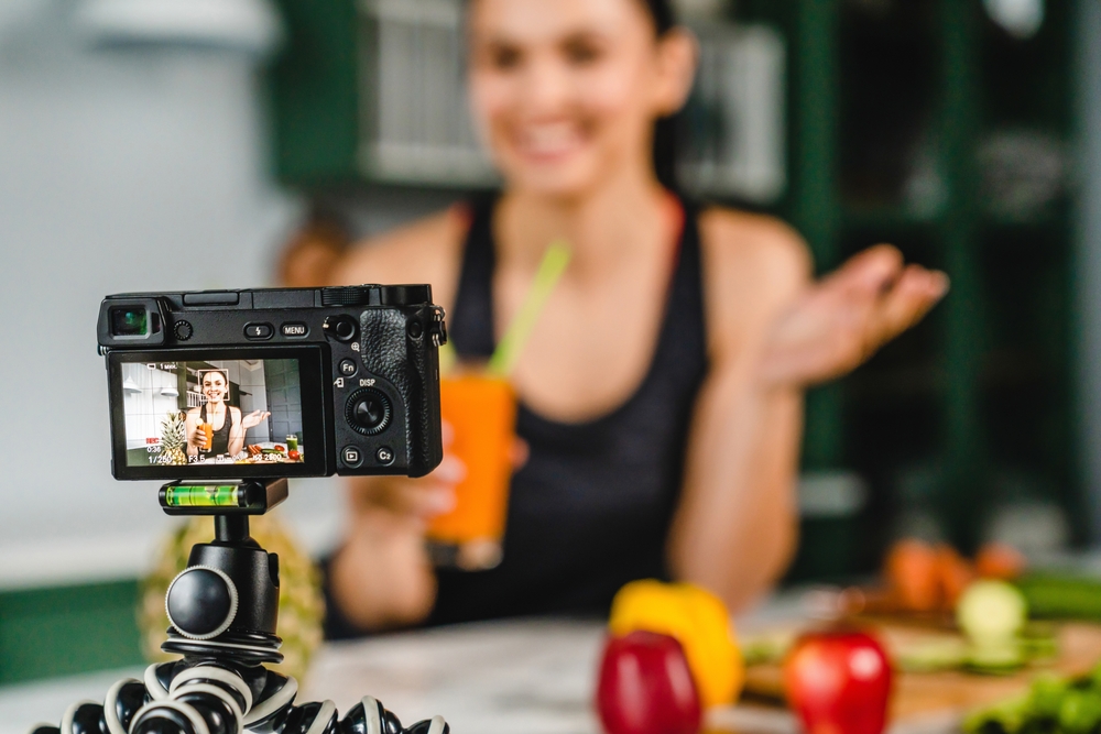 A camera on a tripod records a woman presenting a drink, likely creating content in a kitchen setting.