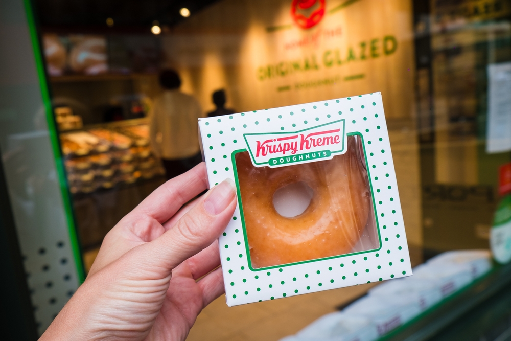A hand holding a single-boxed Krispy Kreme doughnut.