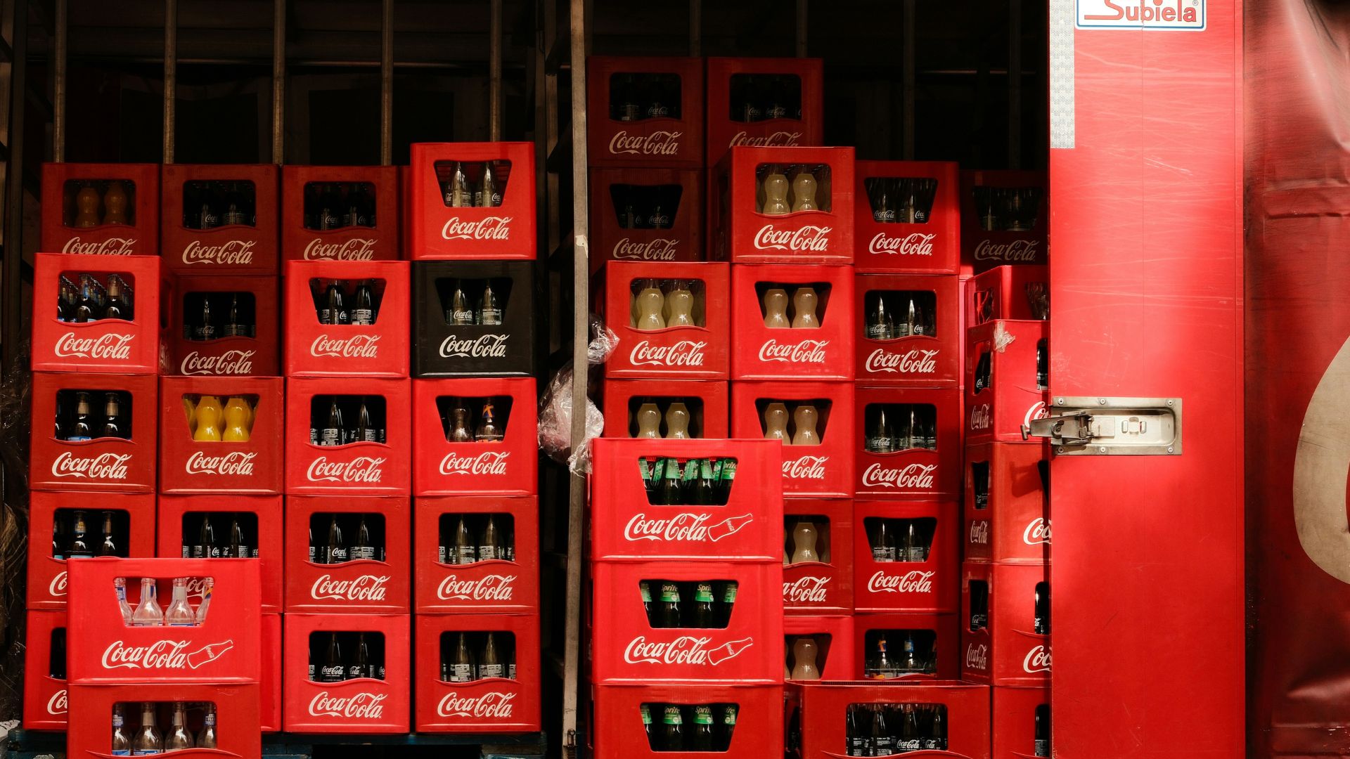 Stacks of red Coca-Cola crates filled with glass soda bottles inside a beverage distribution warehouse.