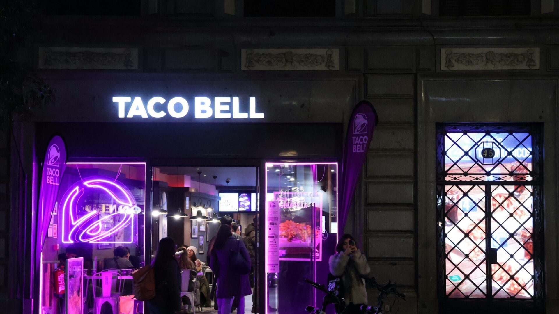 People walking past a brightly lit storefront with purple lighting at night.