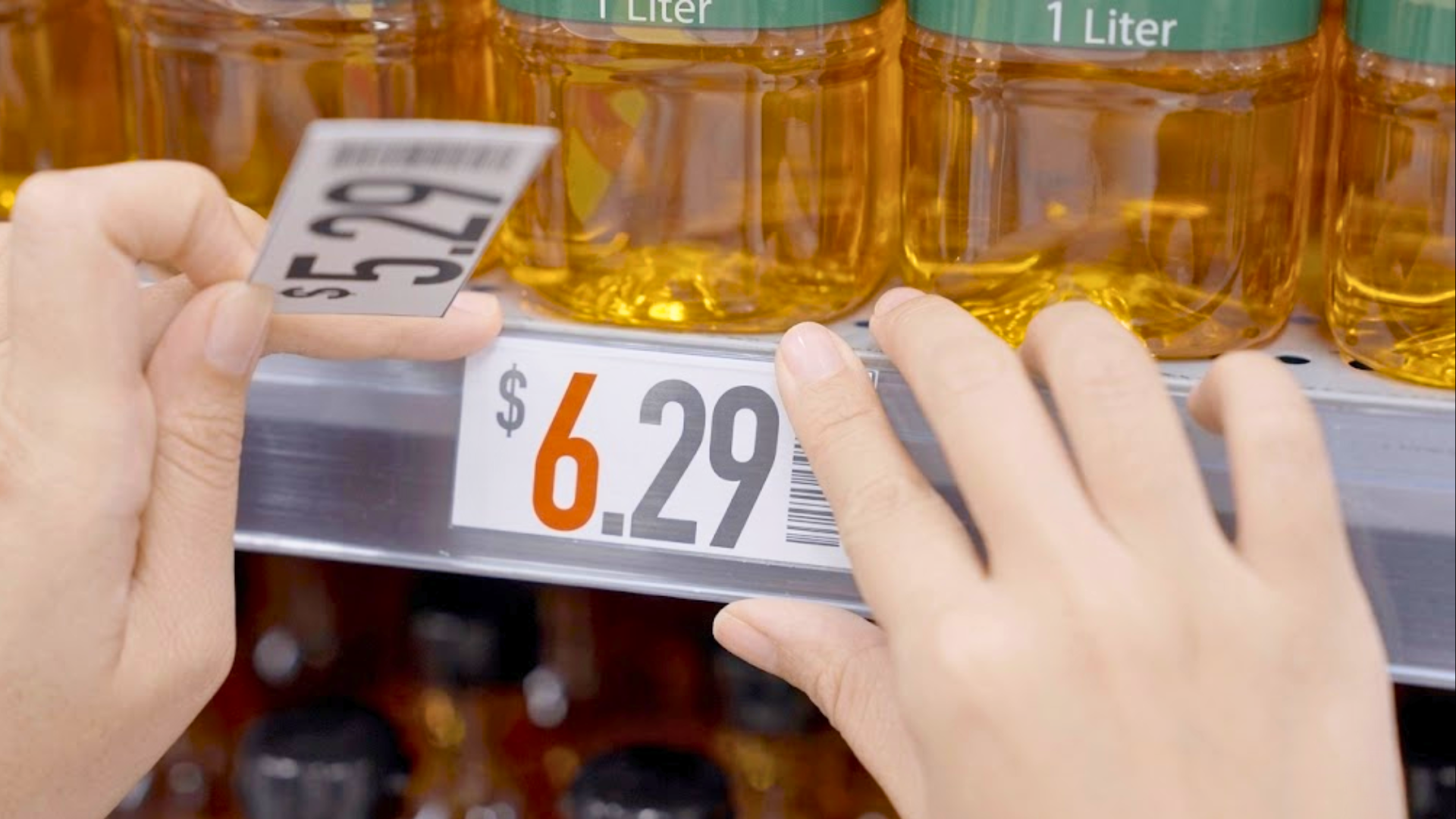 Close-up of hands replacing a $5.29 price tag with a higher $6.29 tag on a grocery shelf stocked with bottles of cooking oil.
