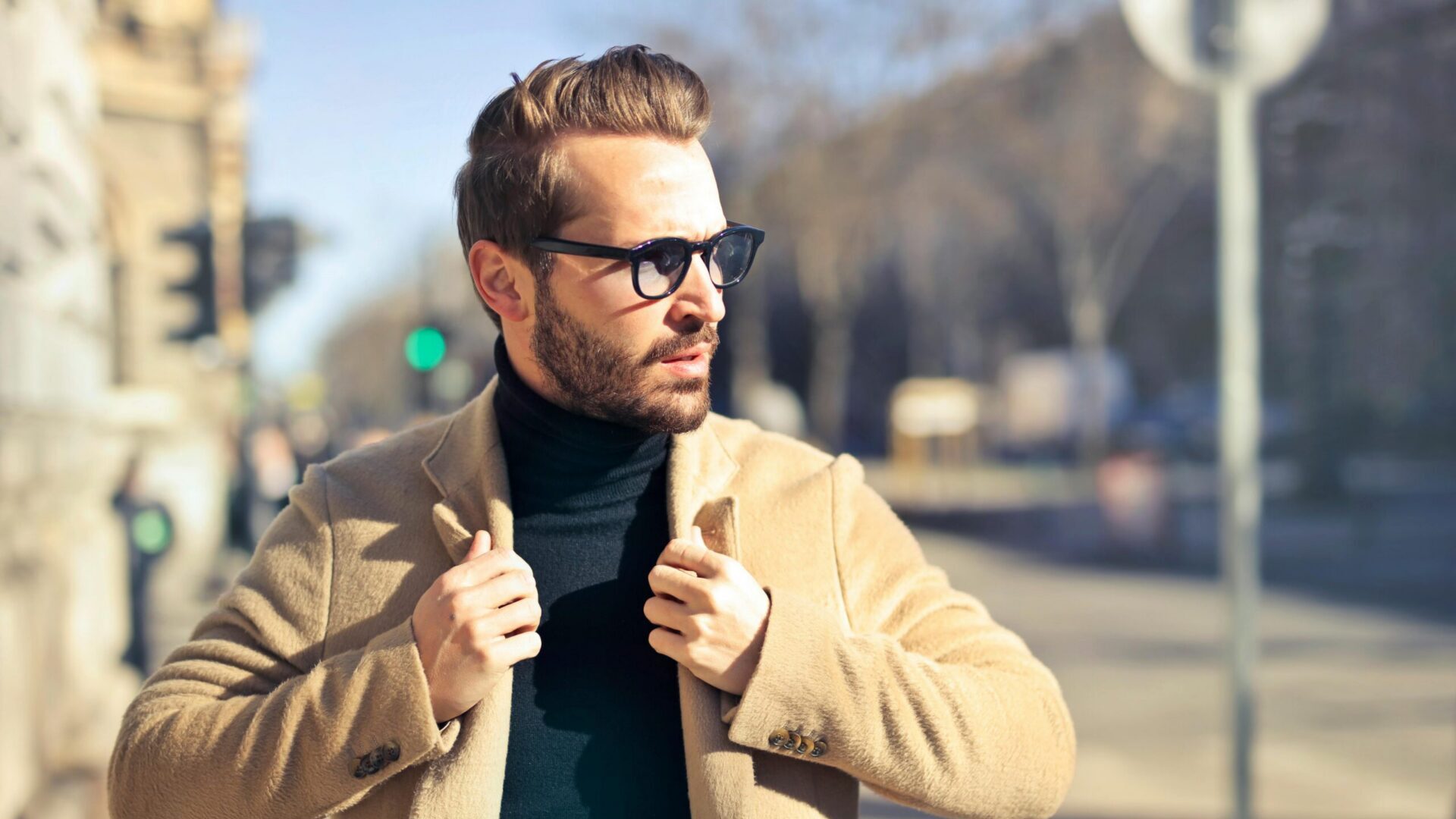 Man wearing sunglasses adjusting his coat while standing outdoors in daylight.