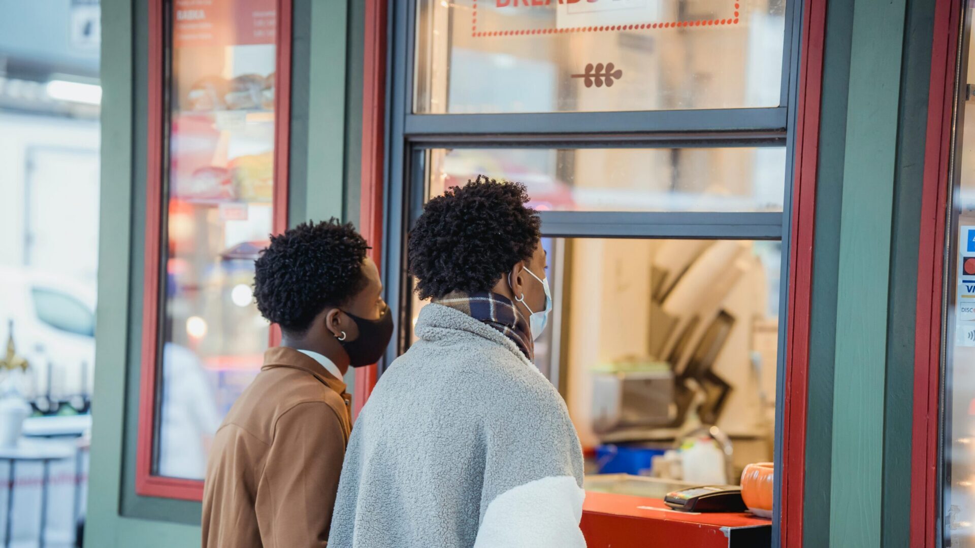 Two men wearing face masks ordering food from a street vendor counter.