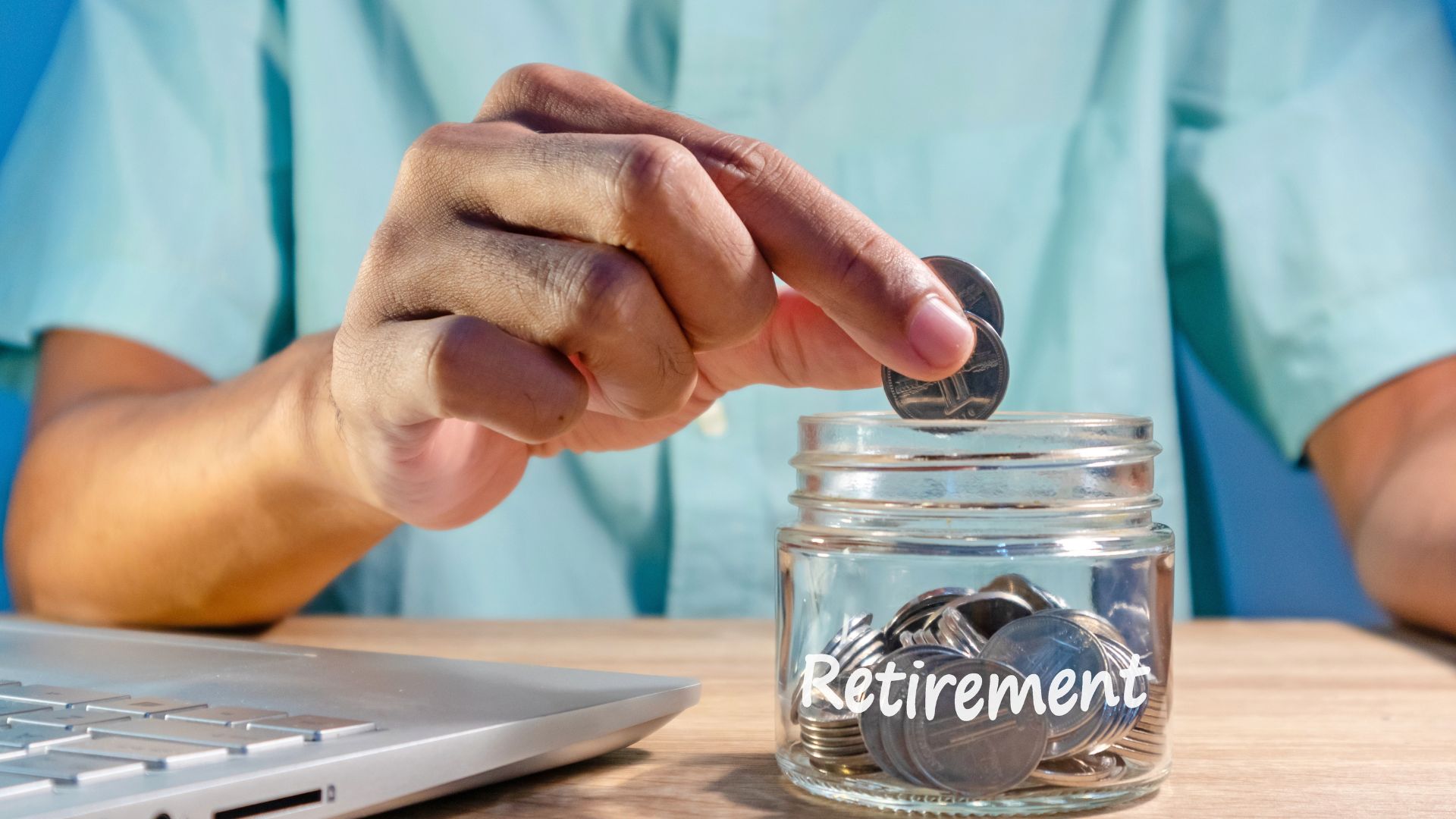 Hand placing a coin into a glass jar labeled retirement next to a laptop.