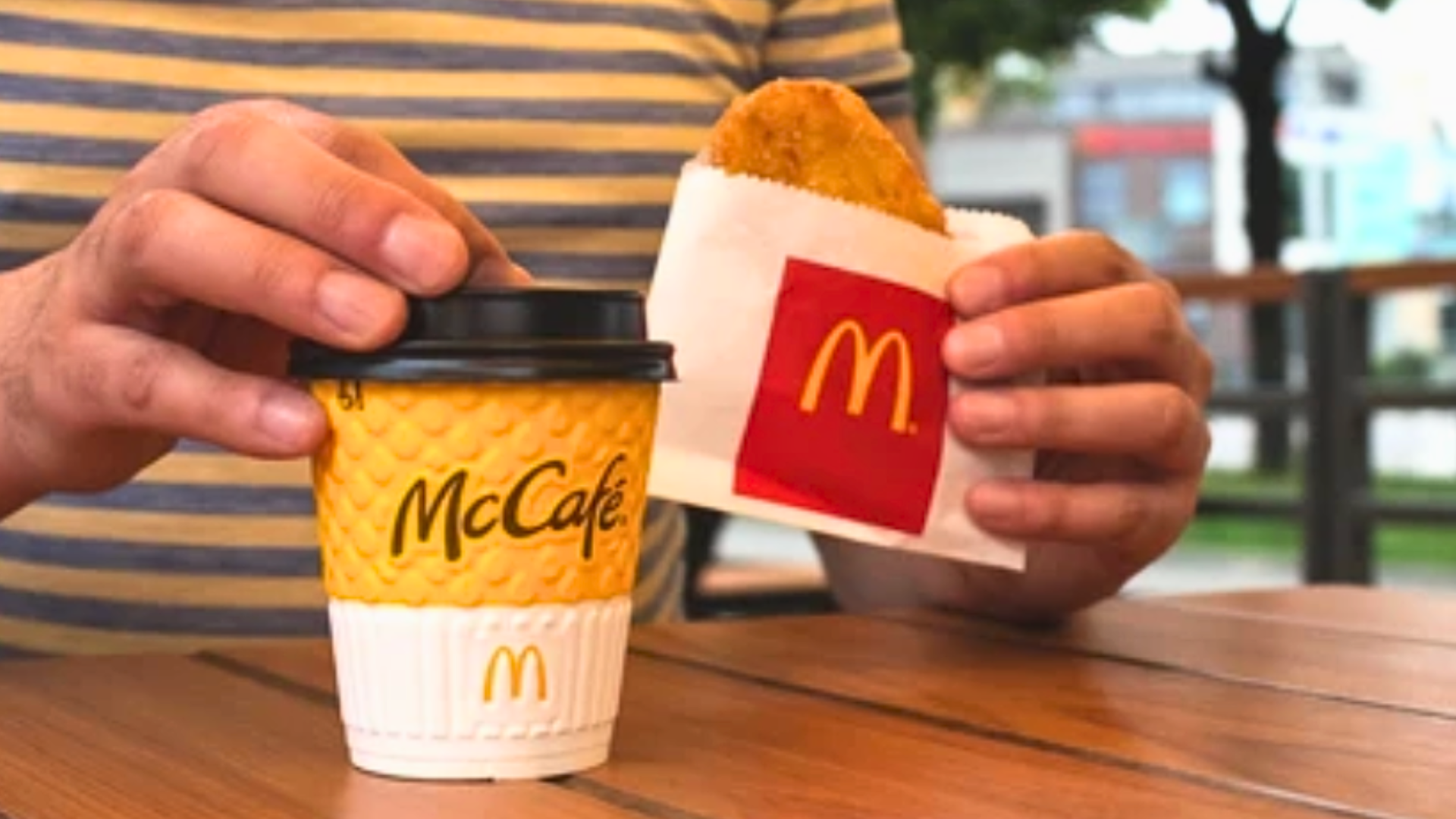 A person holding a yellow McCafé coffee cup in one hand and a McDonald's hash brown in its white and red paper sleeve in the other.