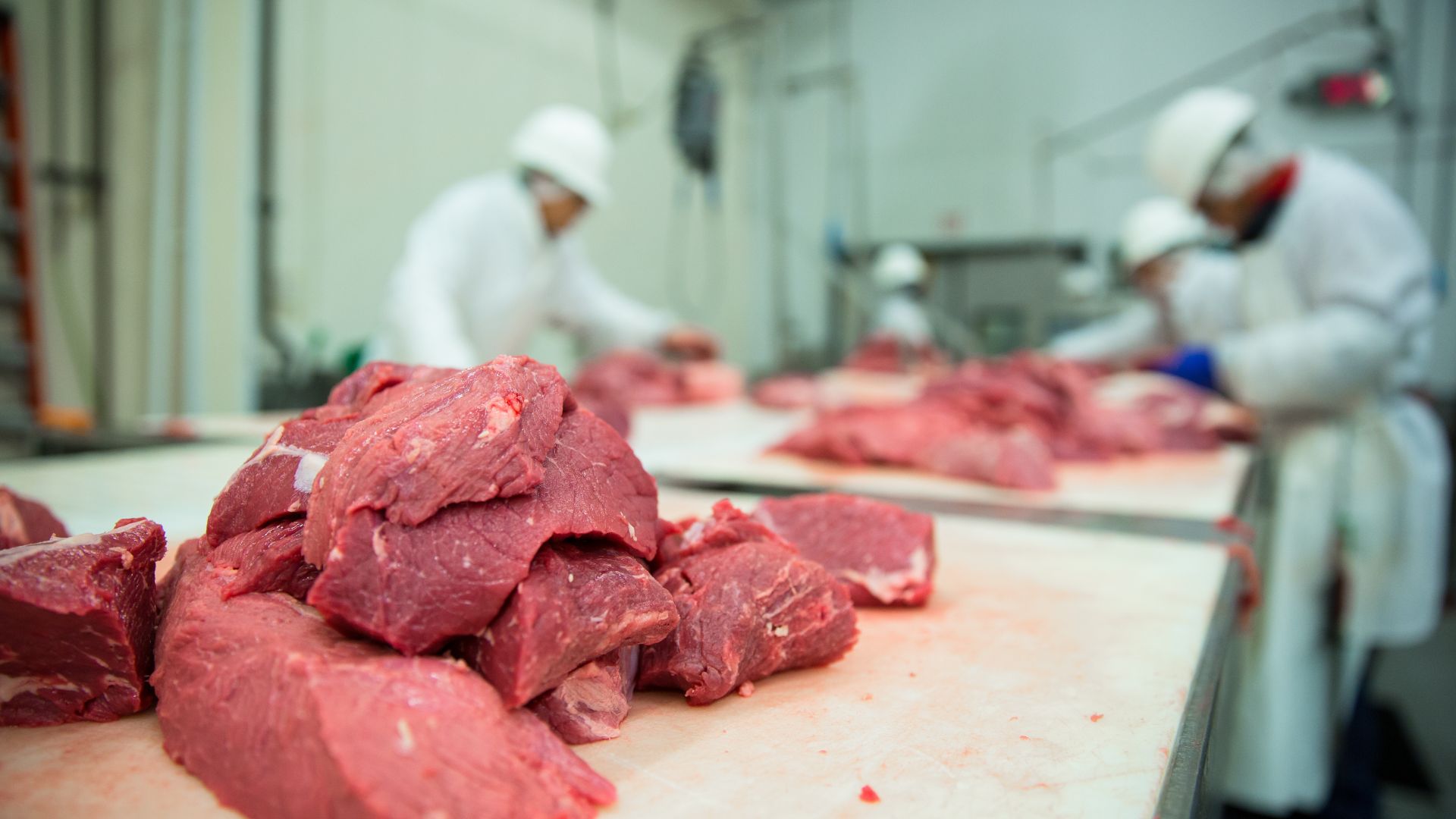 Workers processing raw beef pieces on a table inside a meatpacking plant facility.