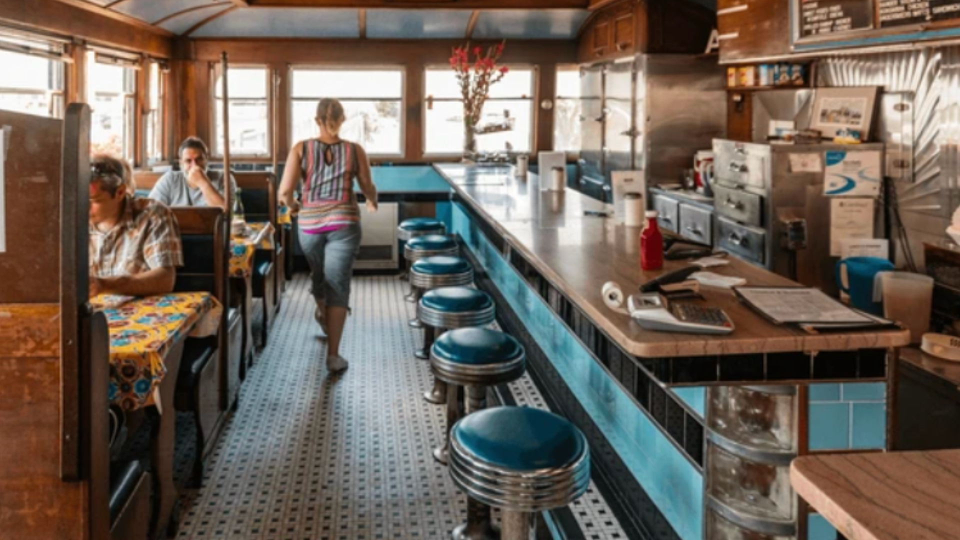 Interior of a vintage-style diner featuring a long counter with blue stools and customers seated in wooden booths.
