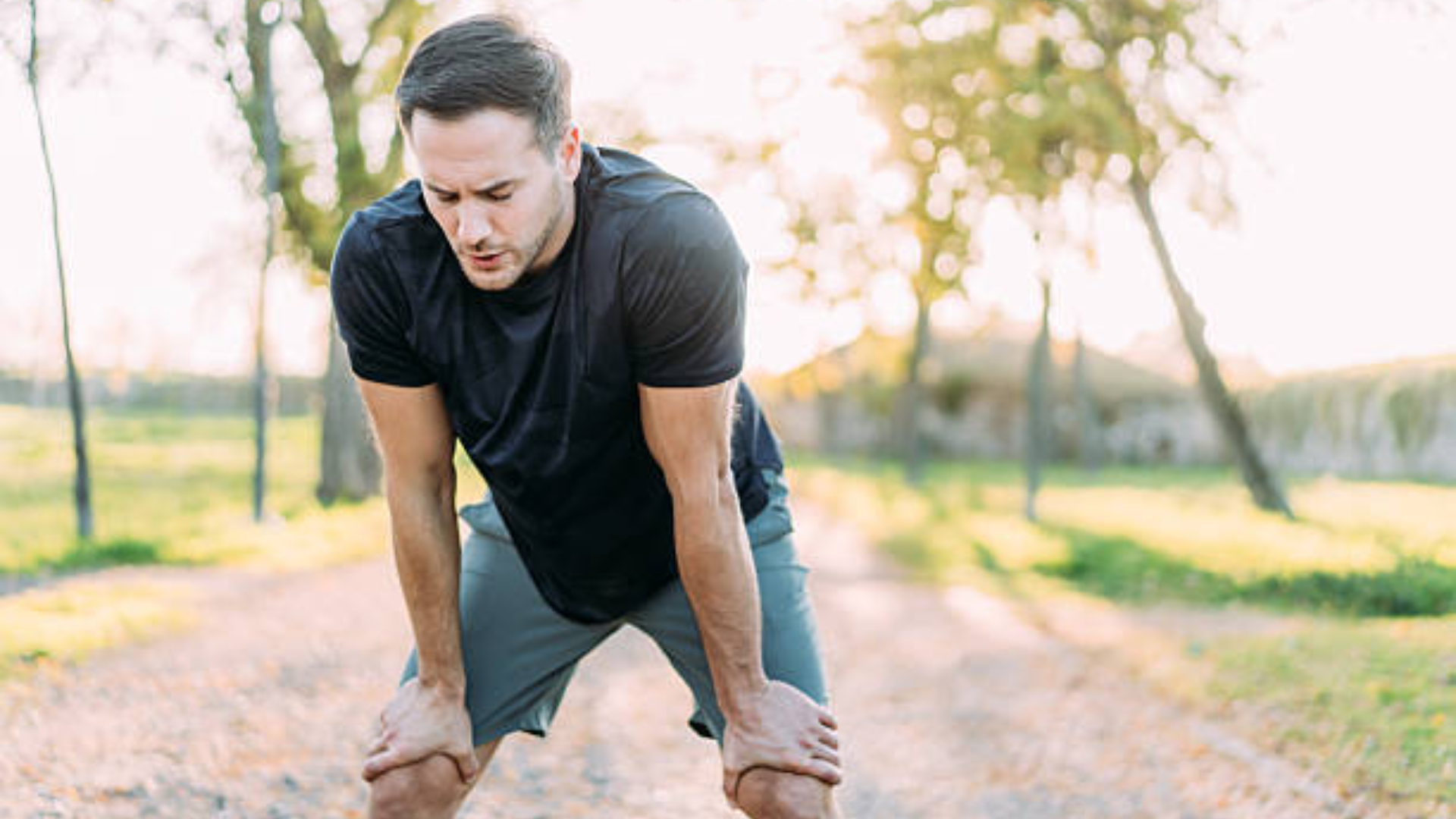 An exhausted male runner in athletic wear leans forward with hands on his knees, catching his breath on a sun-drenched outdoor path.