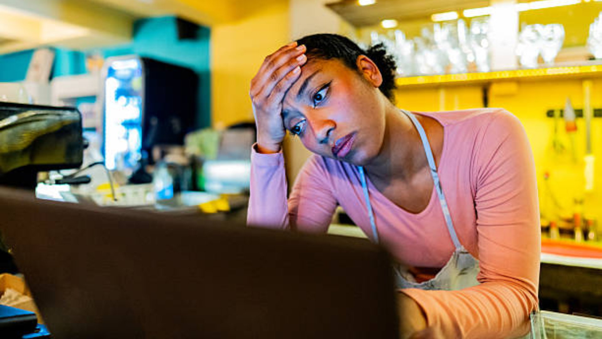A young female fast-food worker in an apron and pink shirt looking stressed and rubbing her forehead while staring at a laptop screen behind a service counter.