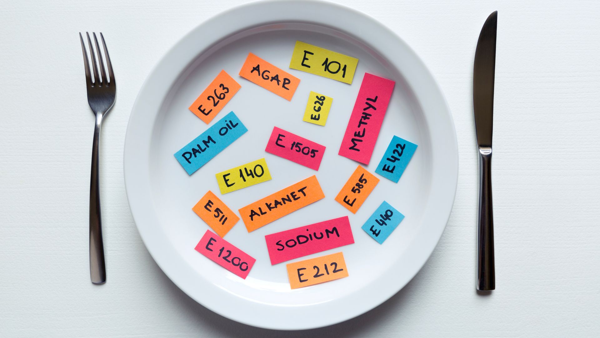 Colorful labels with food additive names and E numbers arranged on a white plate with fork and knife.