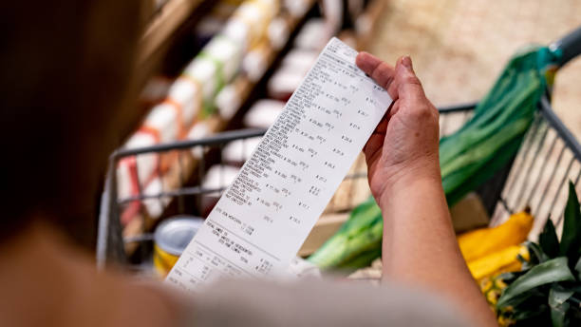 A person’s hand holding a very long grocery store receipt over a shopping cart filled with fresh produce.