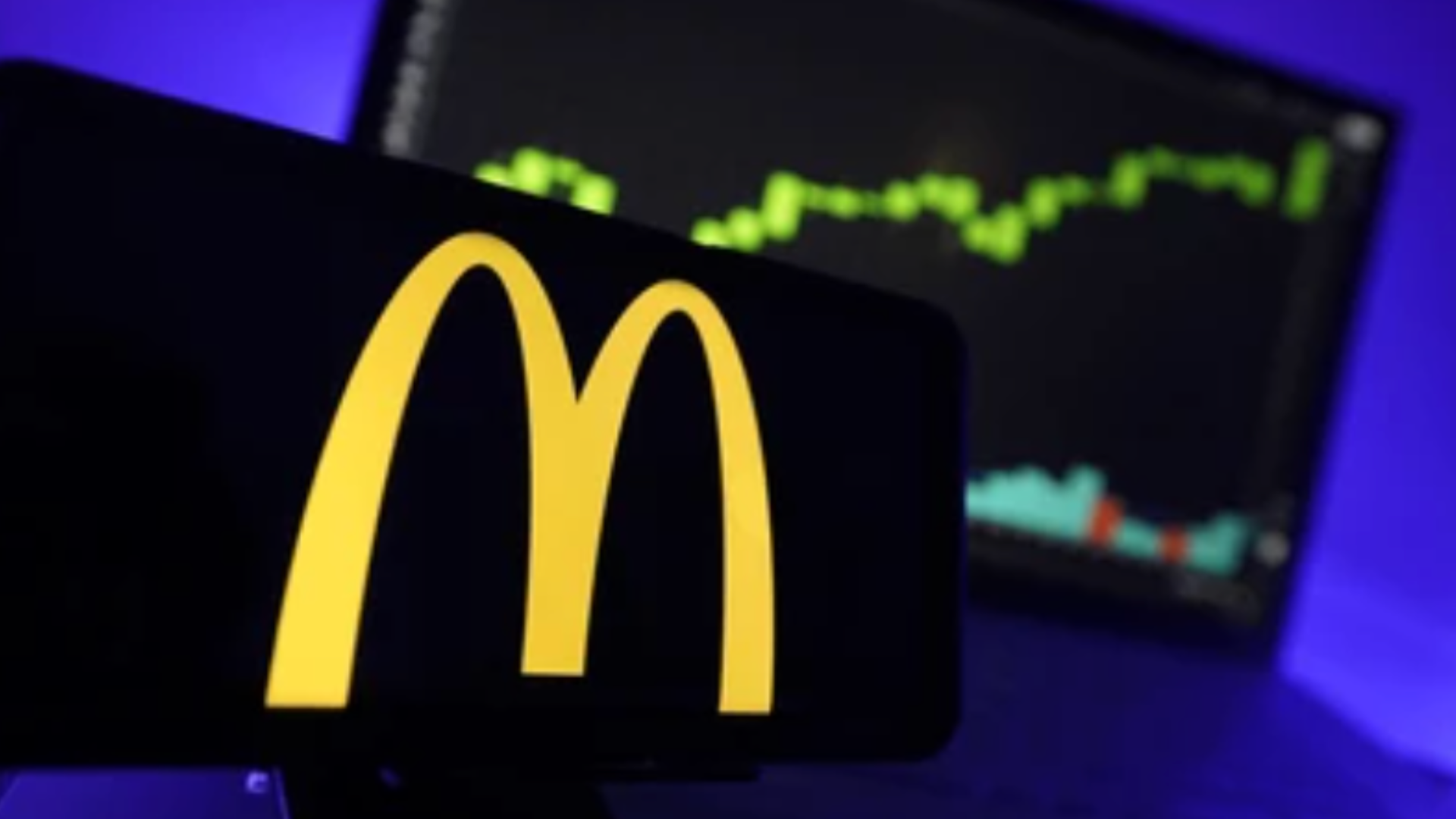 A smartphone displaying the golden arches McDonald's logo in the foreground with a blurred financial stock market graph in the background.
