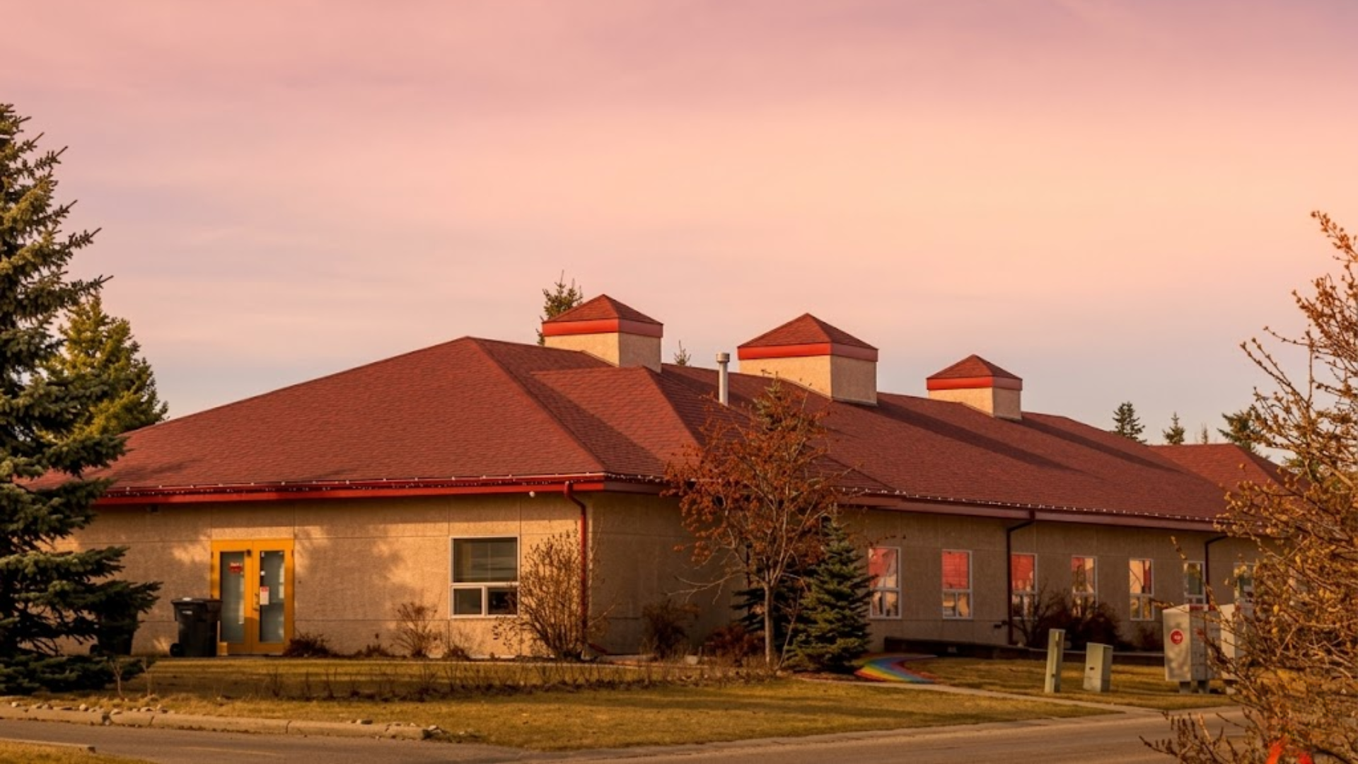 Exterior view of a modern, one-story school building with a large red-shingled roof under a soft sunset sky.