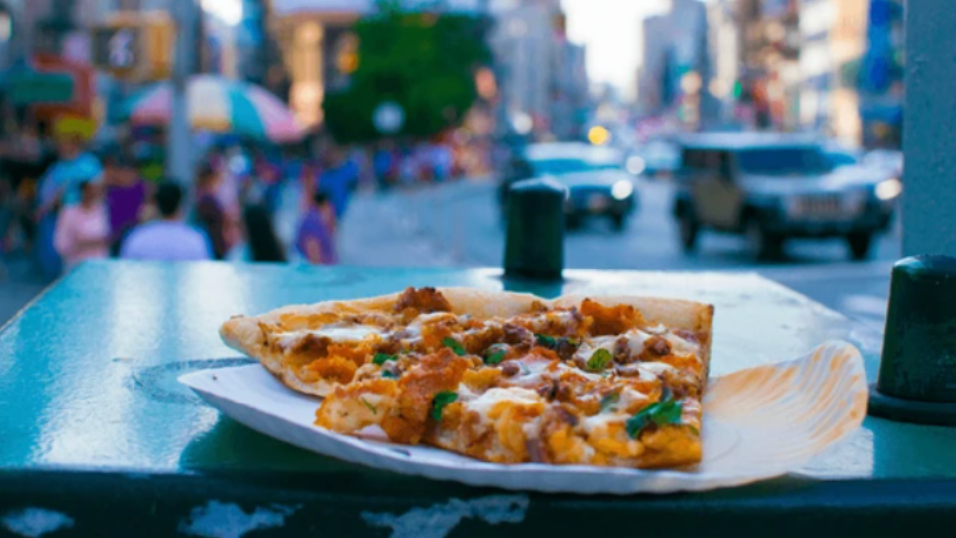 A slice of pizza on a paper plate sitting on a ledge with a blurred street in the background.