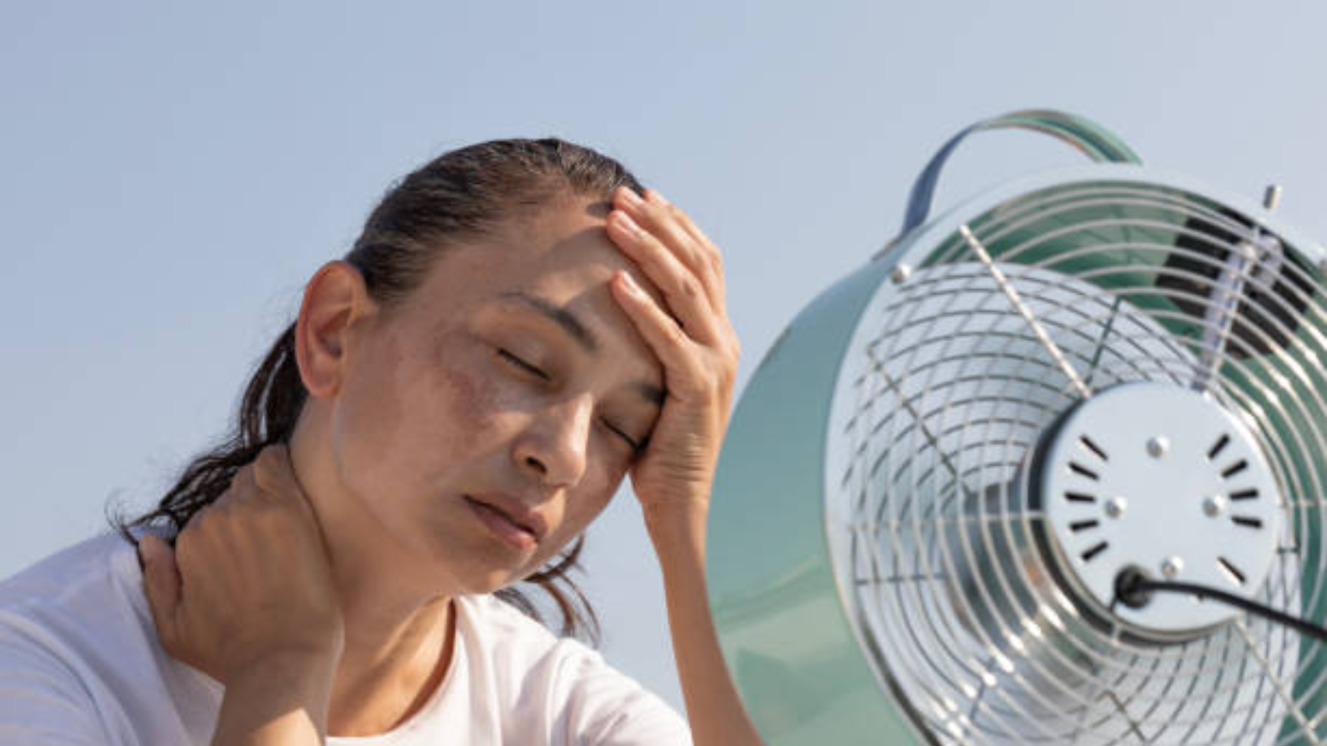 A woman suffering from heat exhaustion holds her head and neck while sitting in front of an electric cooling fan outdoors.