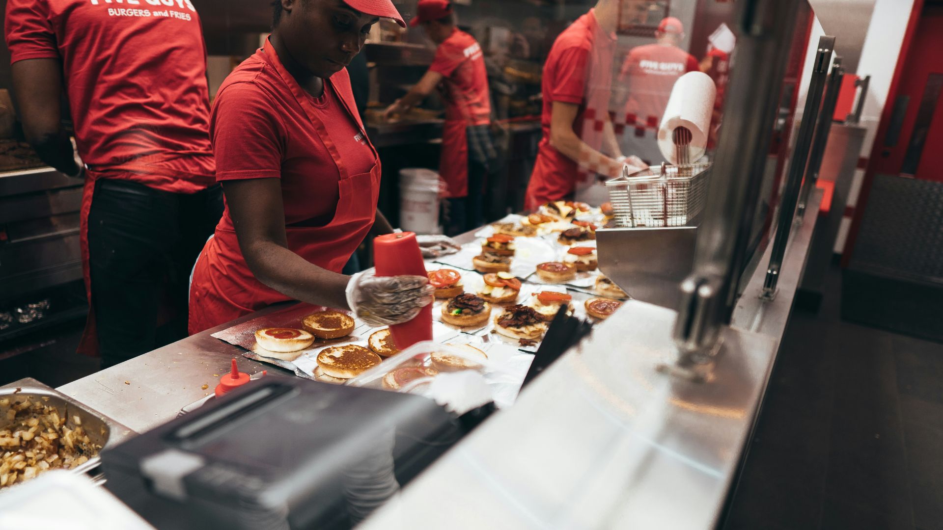 Five Guys employee preparing burgers at a busy kitchen counter inside the restaurant.