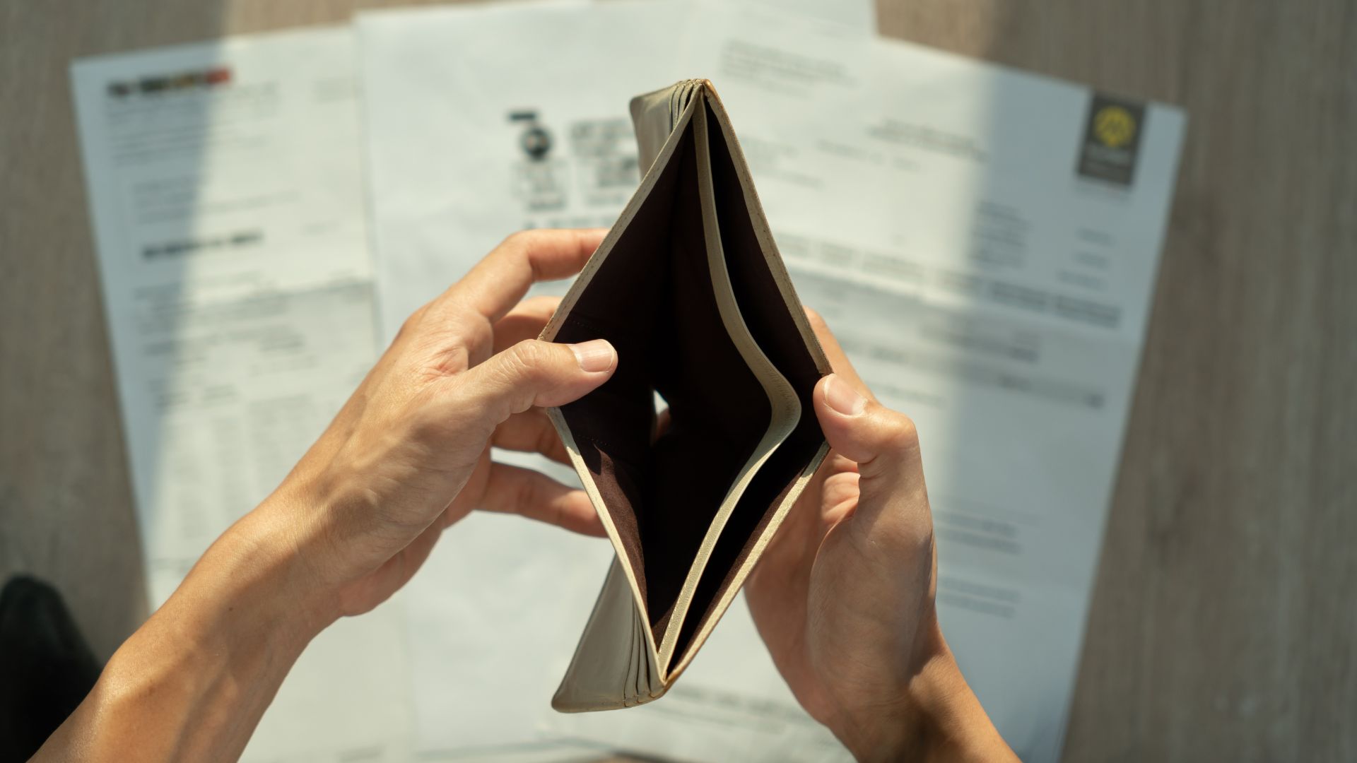 Person holding open an empty wallet with bills and documents blurred in the background.