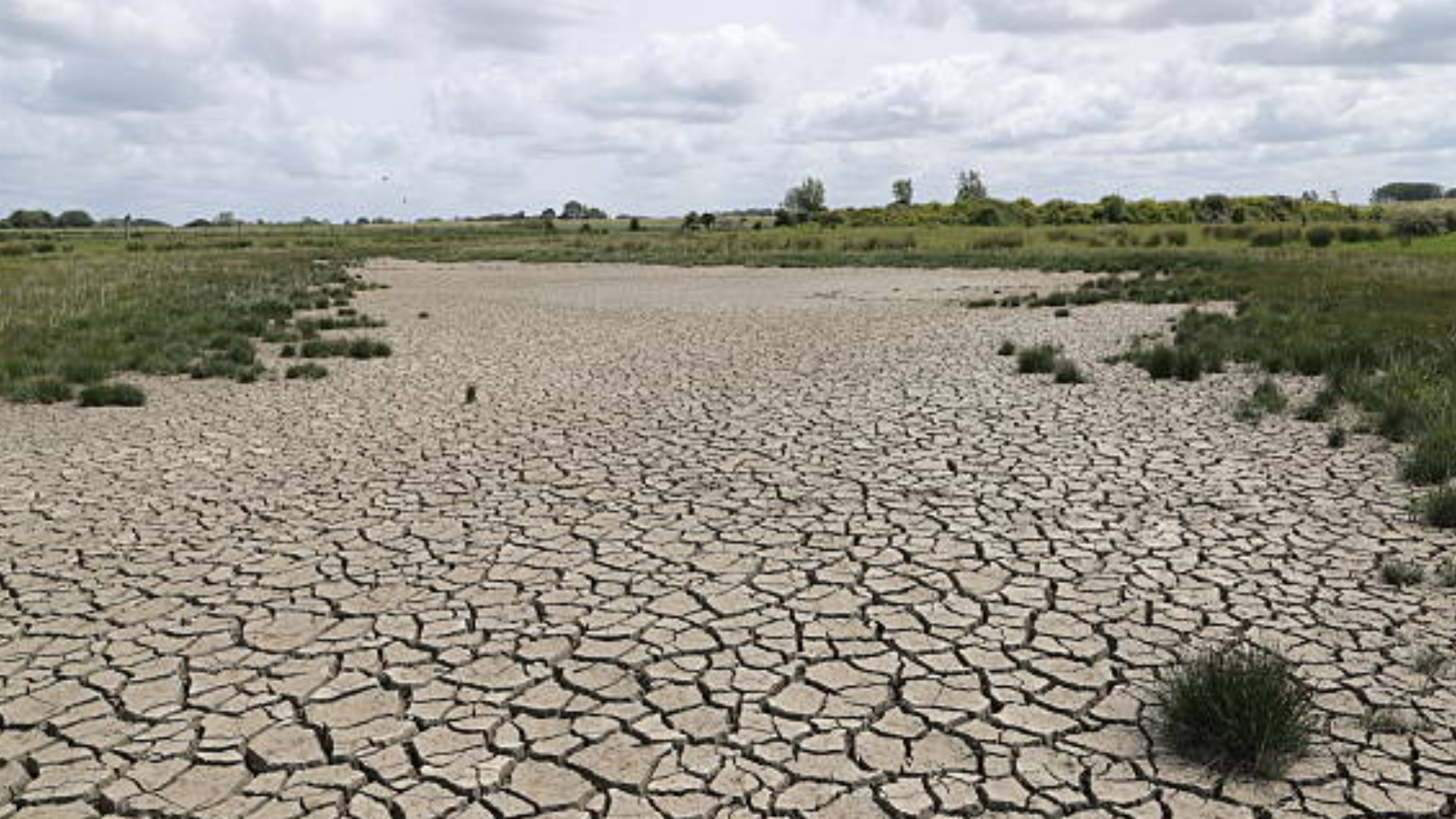 A wide shot of a severely dried-up lake bed with cracked earth under a cloudy sky, indicating drought.