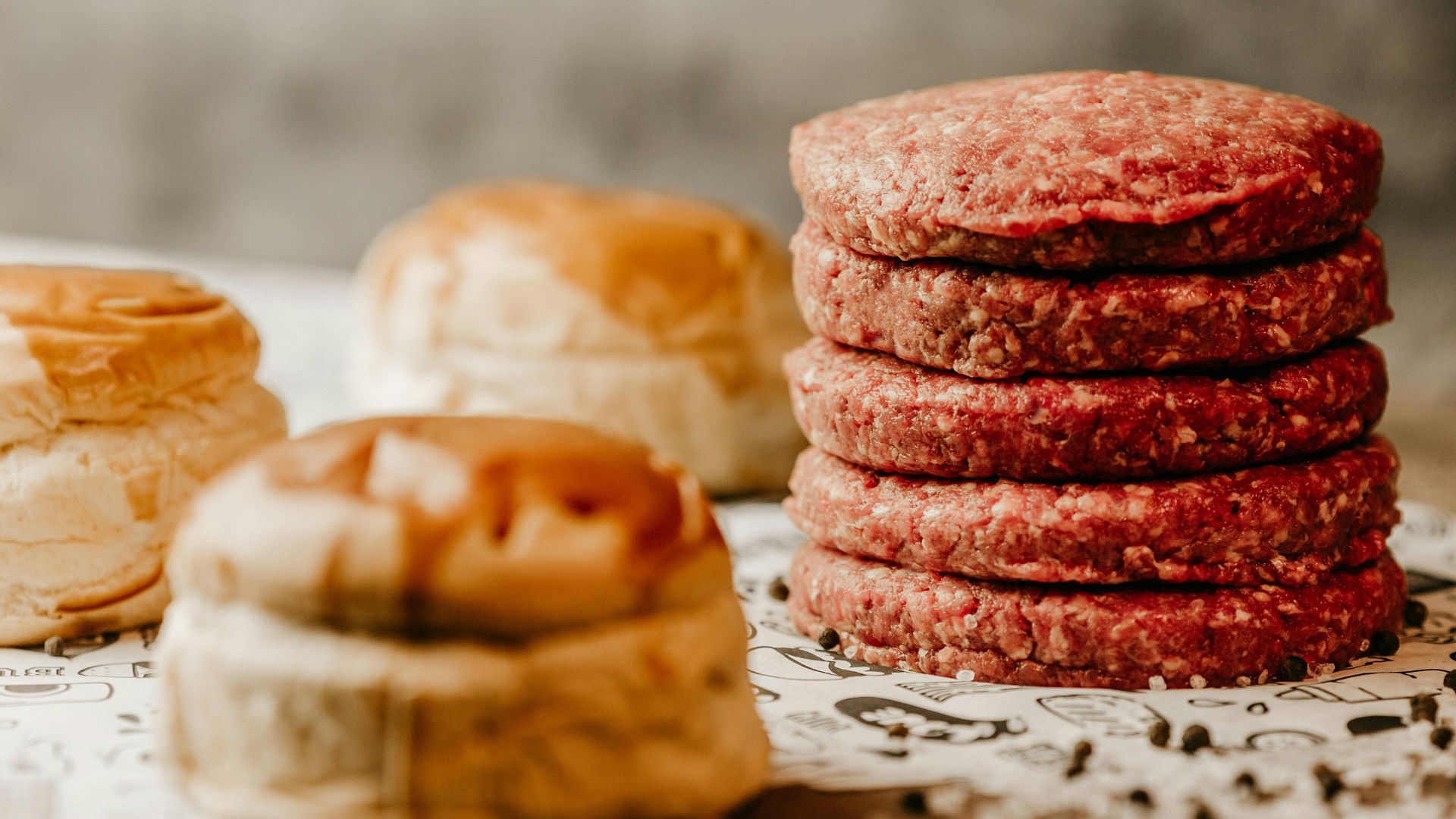 Stack of raw burger patties next to hamburger buns on a prep surface.