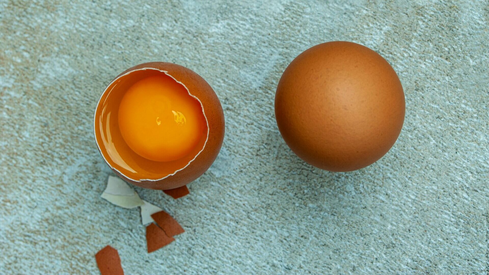 Cracked egg with yolk in shell beside a whole brown egg on a surface.