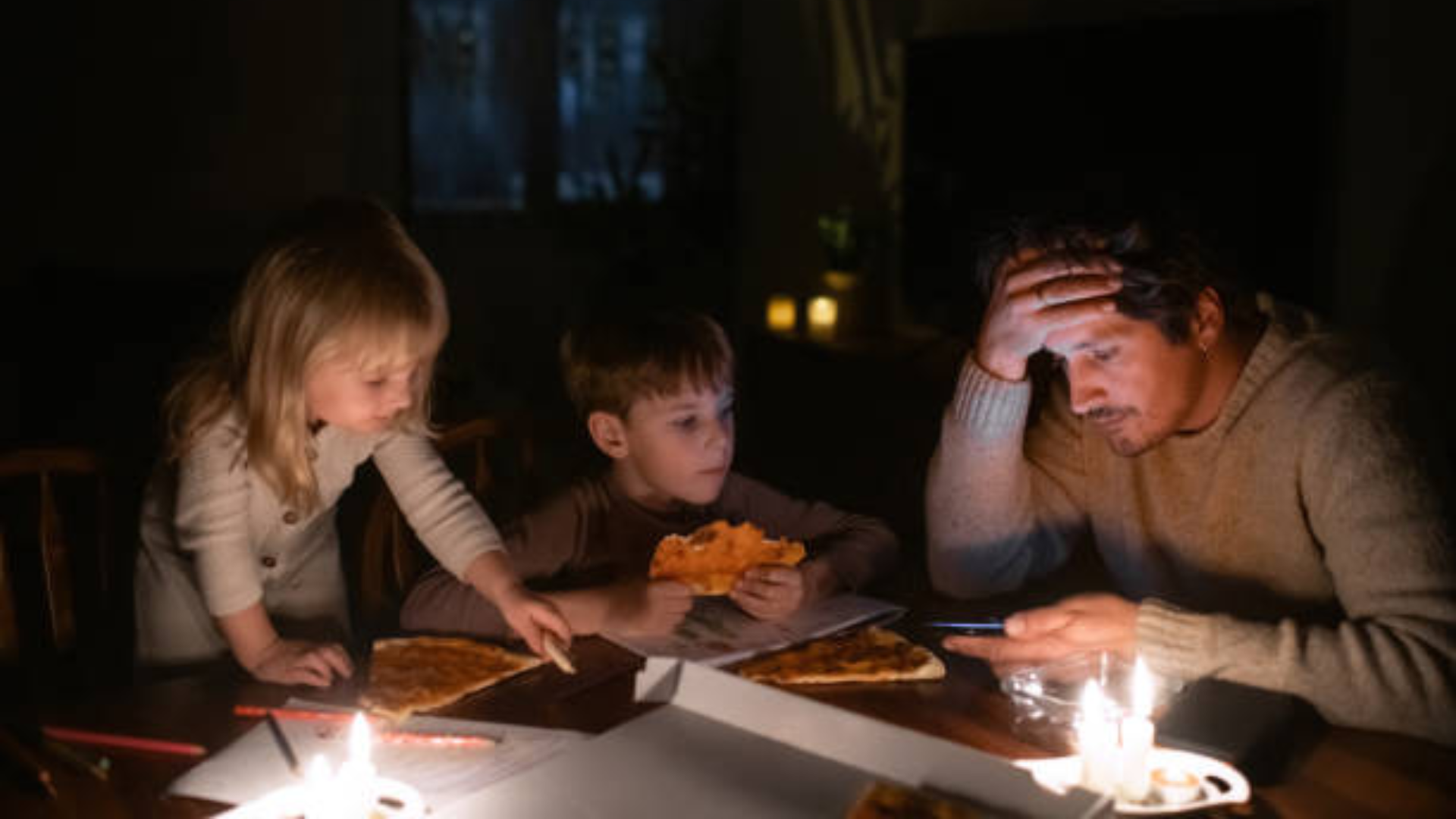 A family eating pizza by candlelight in a dark room, with a man looking stressed while checking his phone.