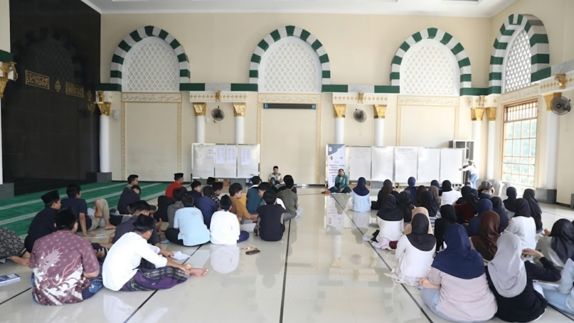 A group of students sits on the floor of a bright, arched hall with Islamic architectural details, listening to a presentation.