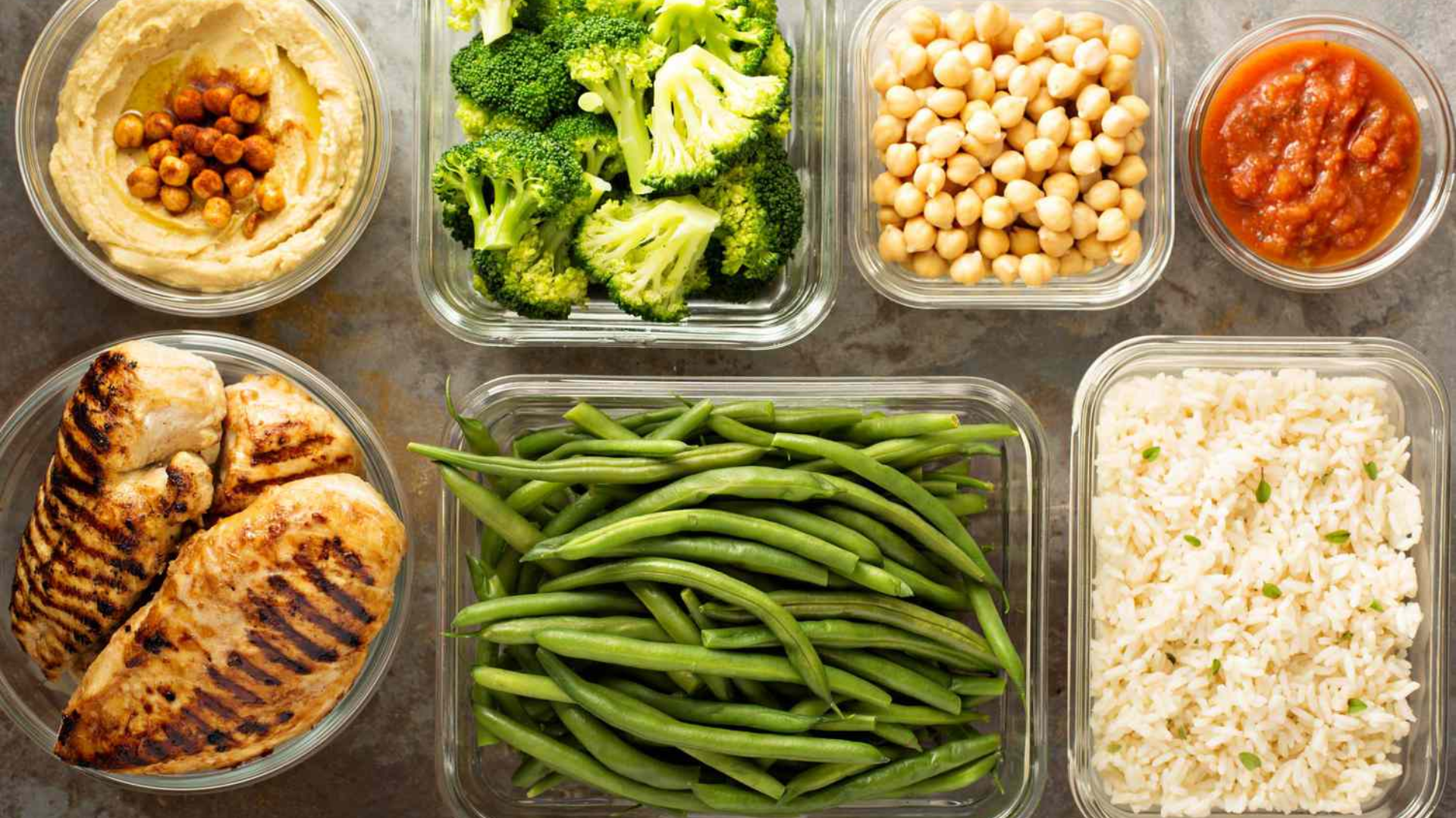 An overhead view of healthy meal prep containers filled with grilled chicken, green beans, broccoli, white rice, chickpeas, and hummus.