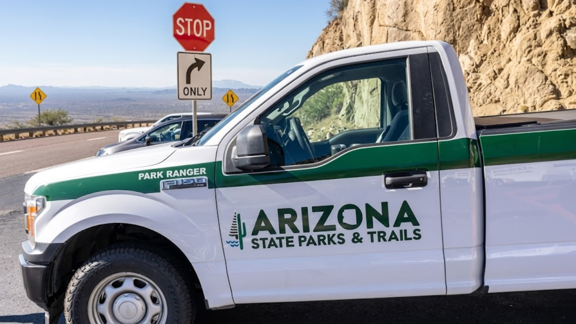 ide view of a white Arizona State Parks & Trails park ranger pickup truck parked on a mountain road overlooking a desert landscape.