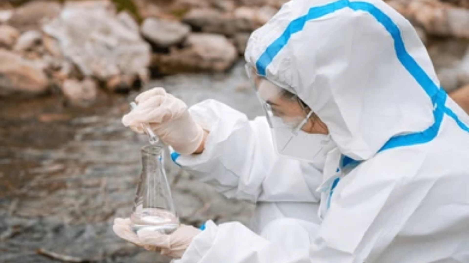 An environmental scientist in a white hazmat suit, mask, and goggles collects a water sample from a rocky river into a glass flask for testing.